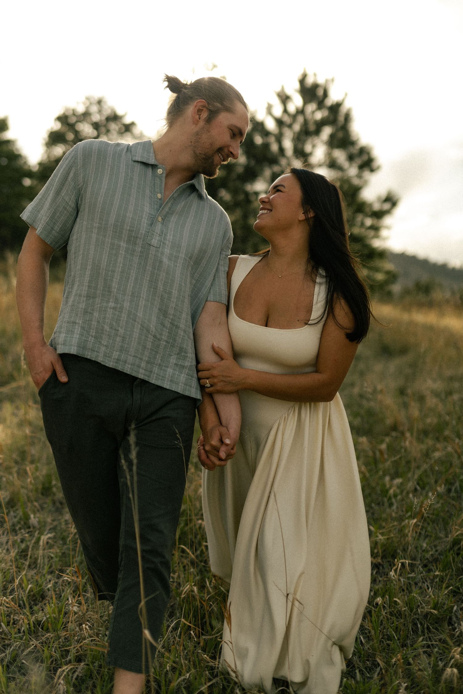 A smiling couple walks hand-in-hand through a golden meadow, gazing at each other at sunset.