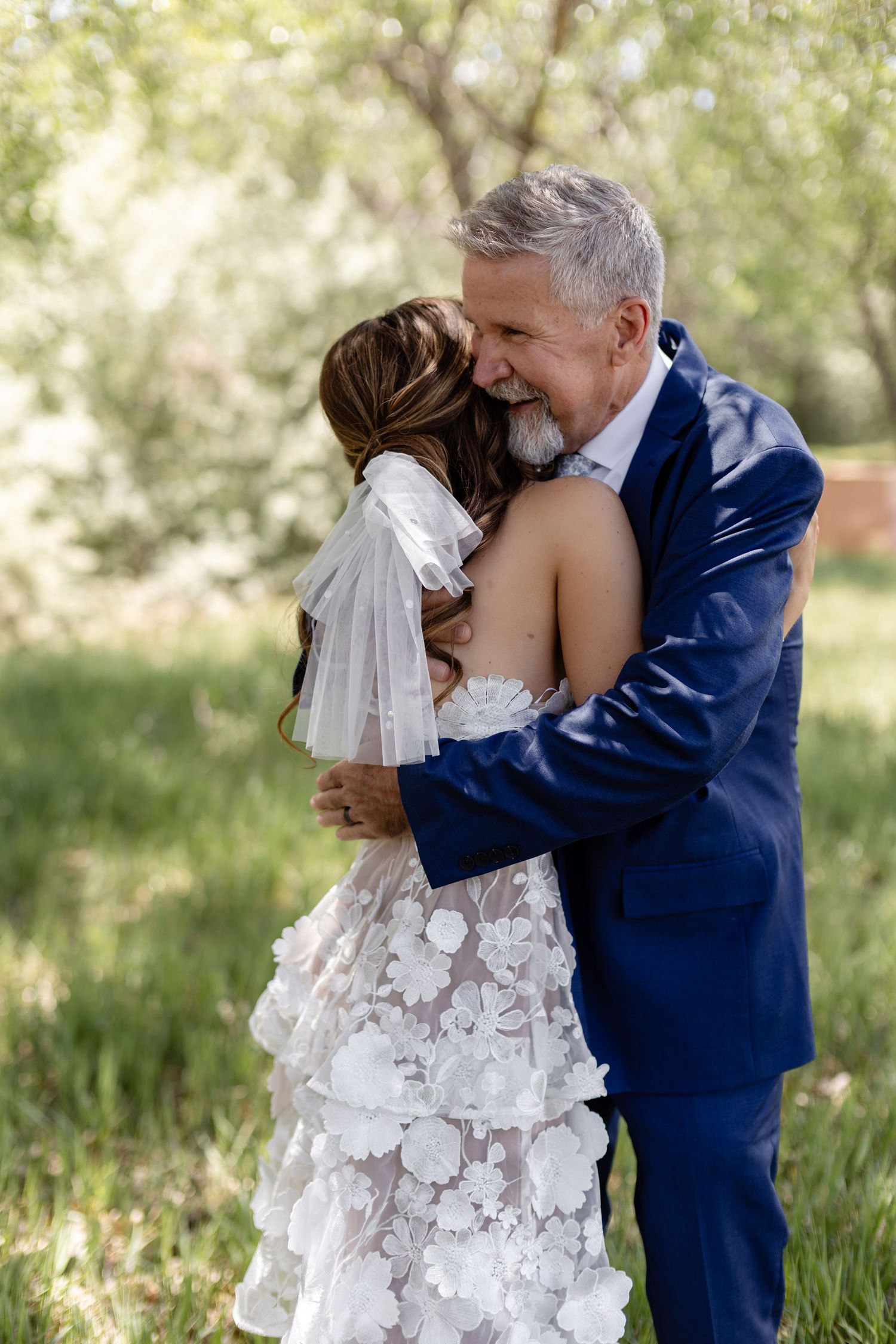 Bride and groom embracing outdoors, man in blue suit hugging woman in floral lace wedding dress with veil.