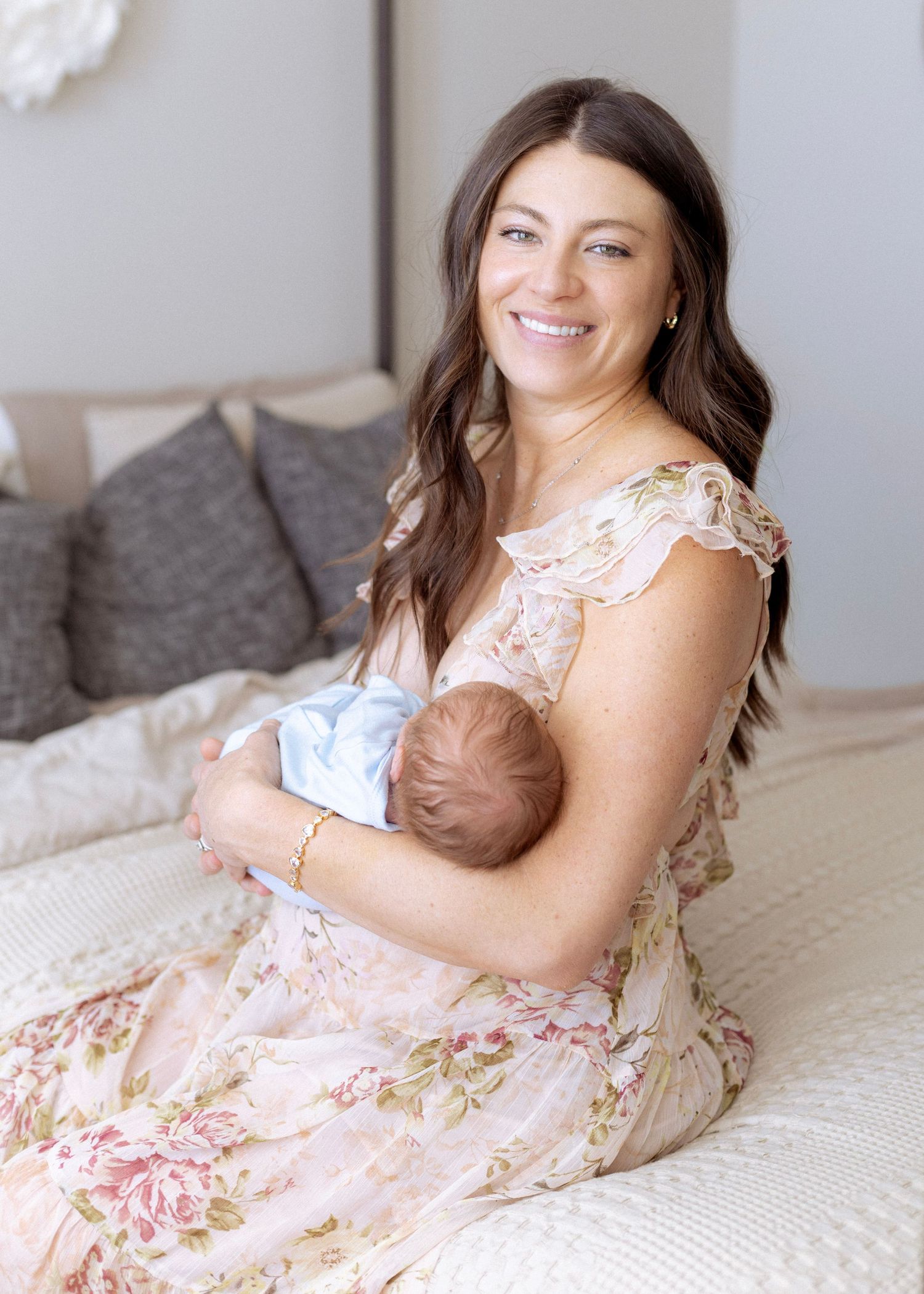Woman in floral dress rests on white bedding while cradling a newborn baby.