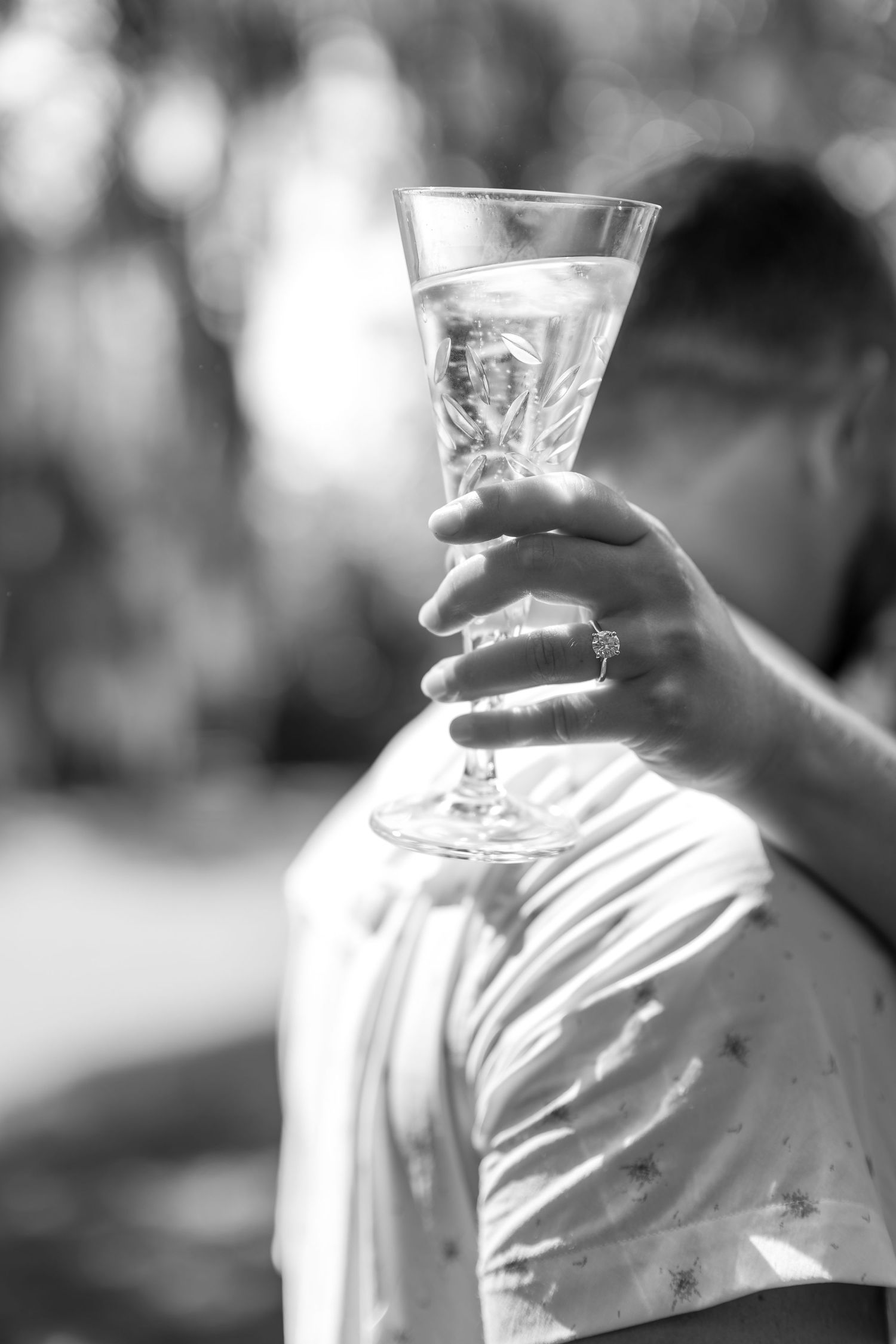Black and white close-up of a hand holding a champagne flute with an engagement ring visible.