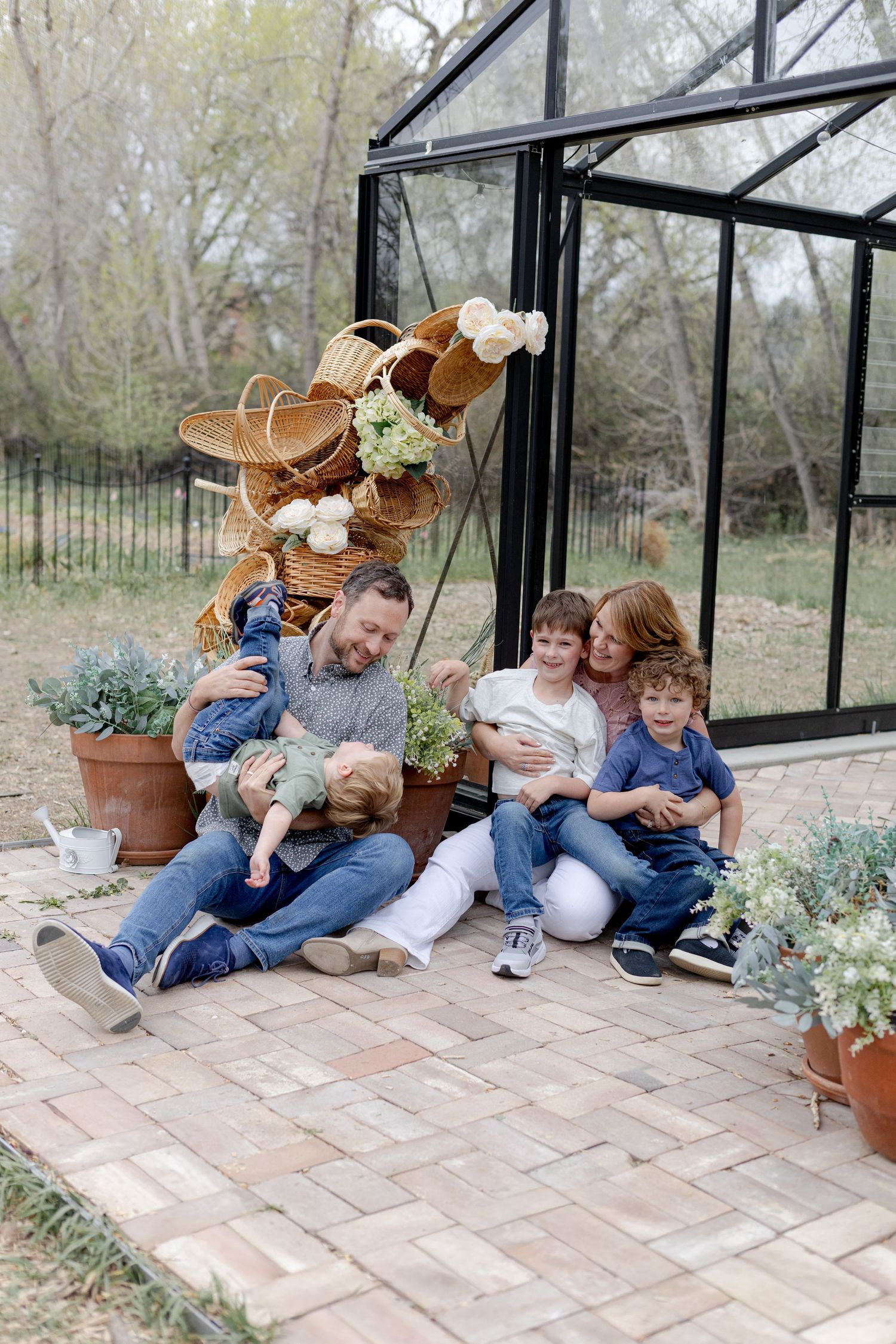 Happy family of five sitting together outside a greenhouse surrounded by wicker baskets and potted plants.