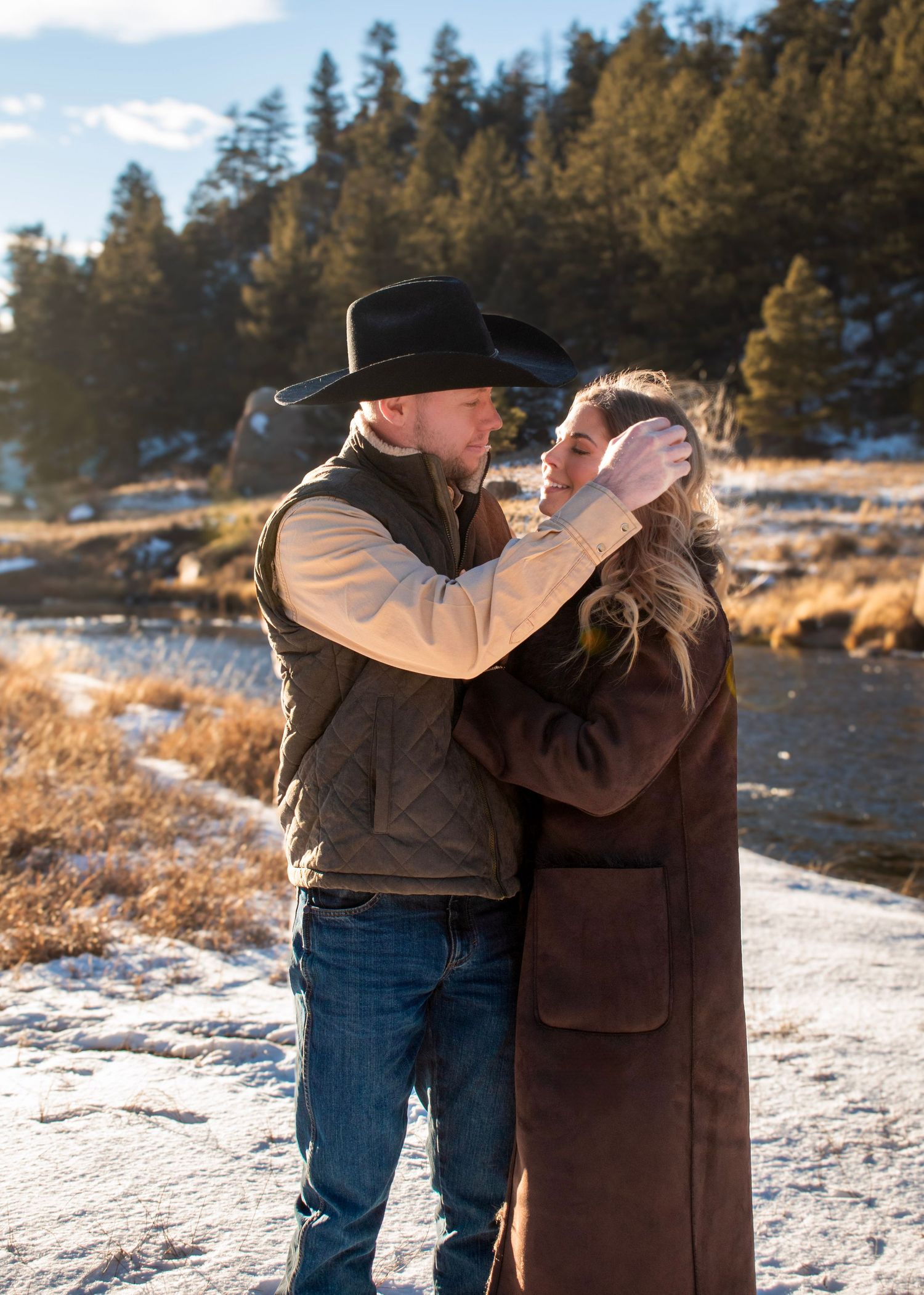 Two people in winter attire embrace in a snowy mountain setting with evergreen trees.
