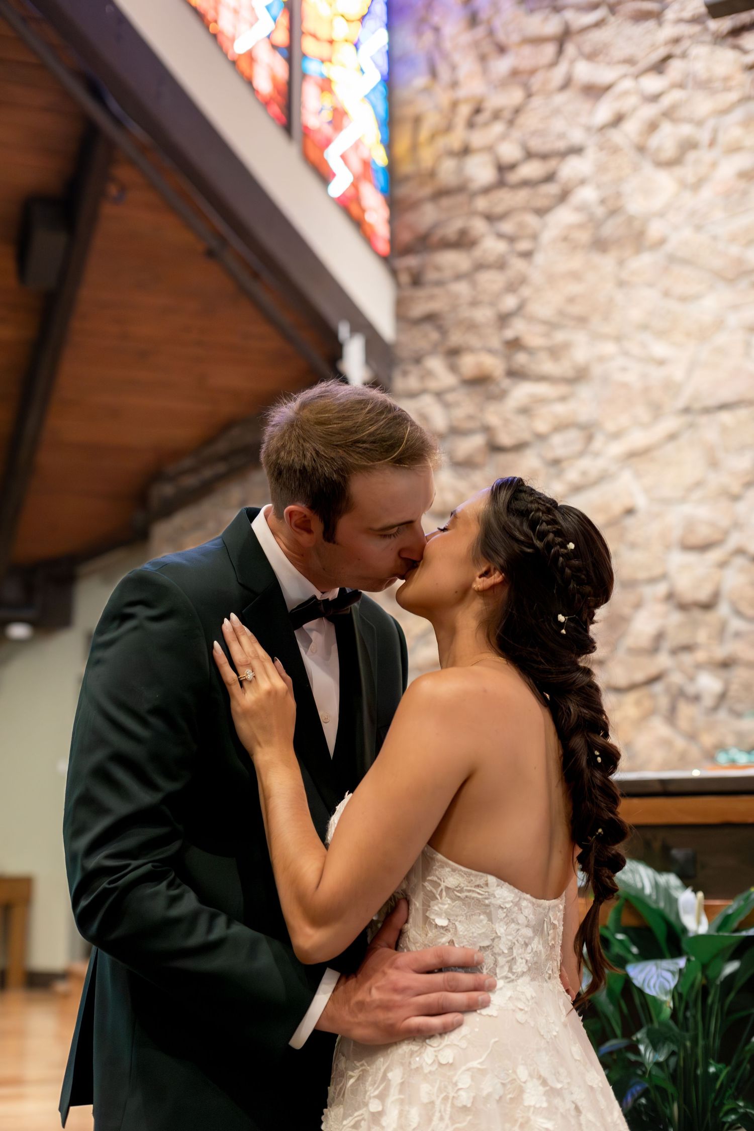 Bride and groom share first kiss at church altar, groom in black tuxedo, bride in lace gown with braided hair.