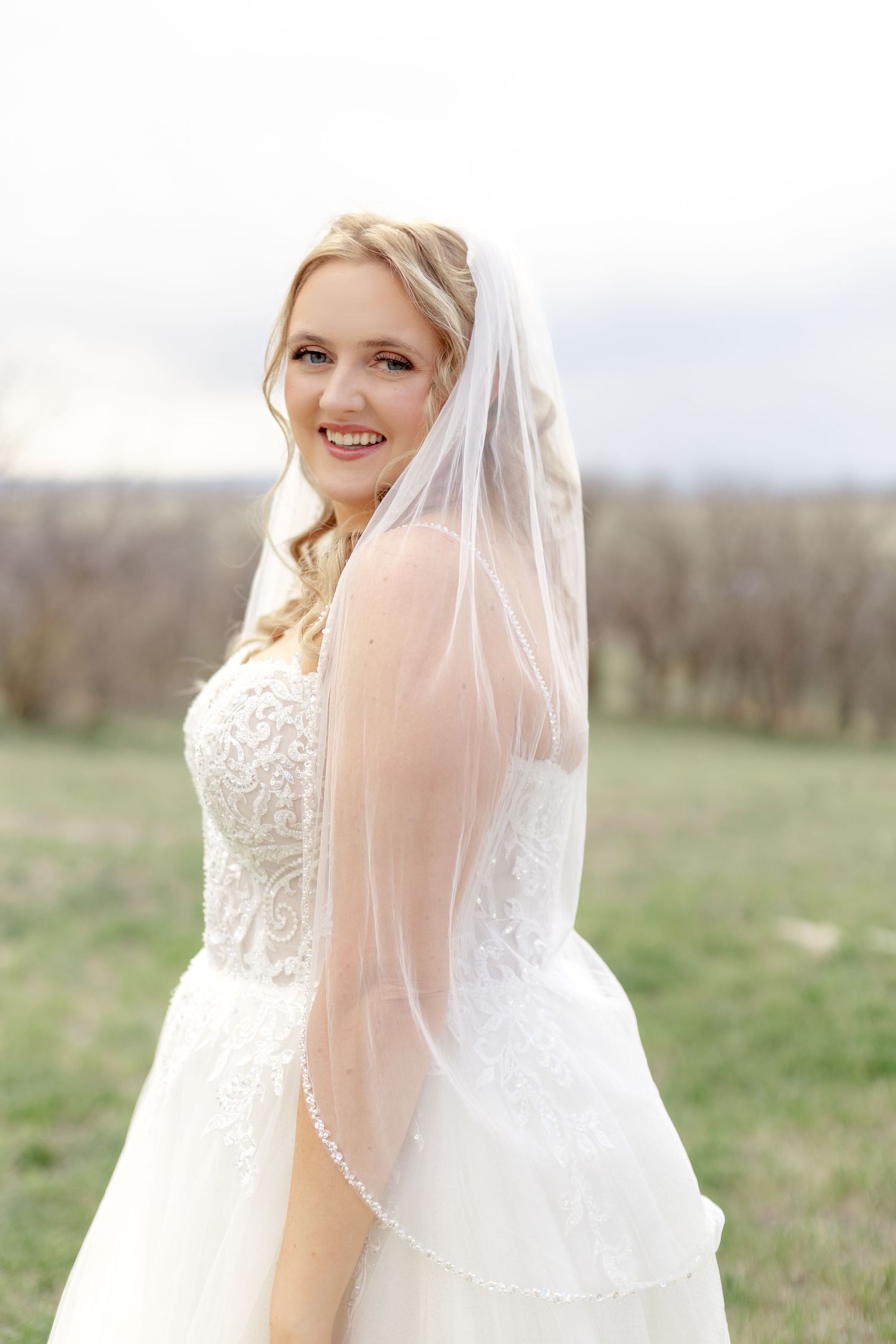Smiling bride in lace wedding gown and pearl-edged veil posing outdoors on wedding day.