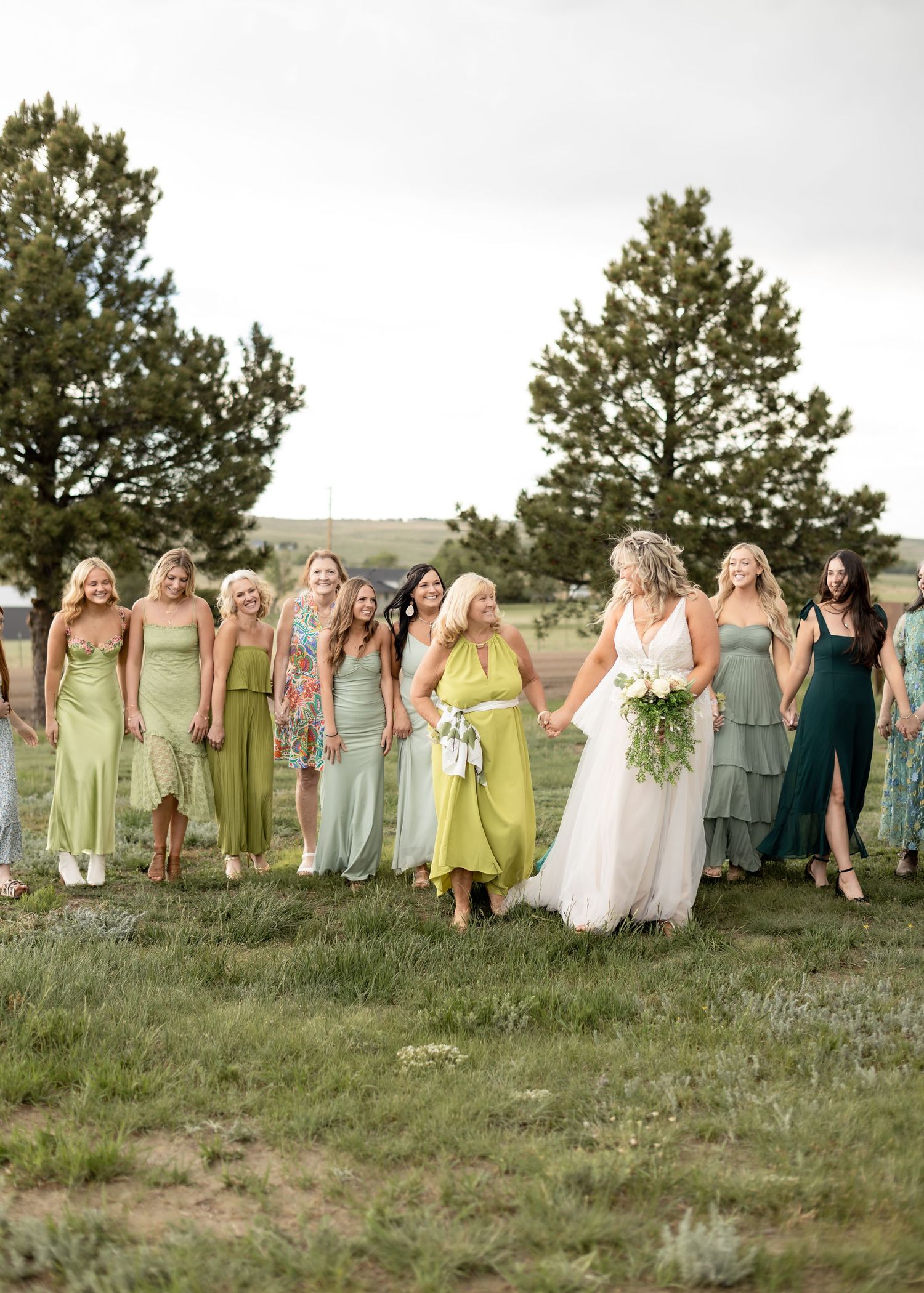 Bride walks with large bridal party in mismatched green dresses across a scenic outdoor field.