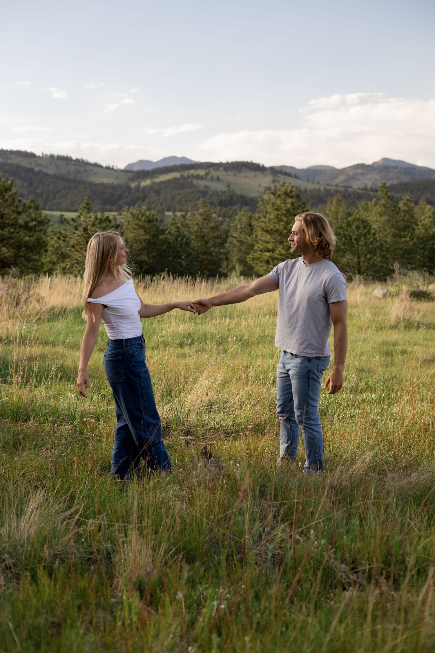 Couple holding hands in a scenic mountain meadow with evergreen trees and rolling hills in the background.