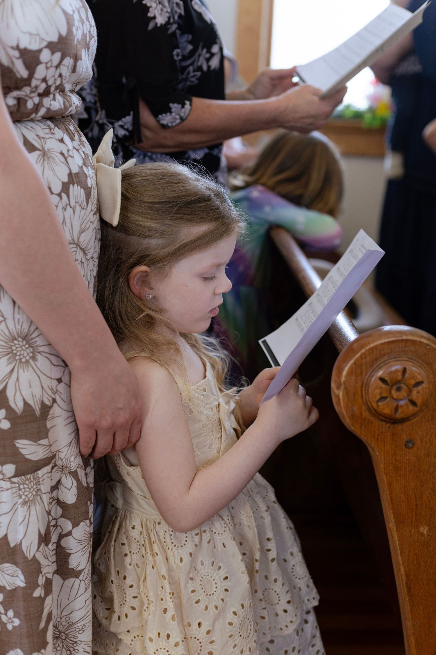 Young girl in white dress reads church program during wedding ceremony beside woman in floral dress.