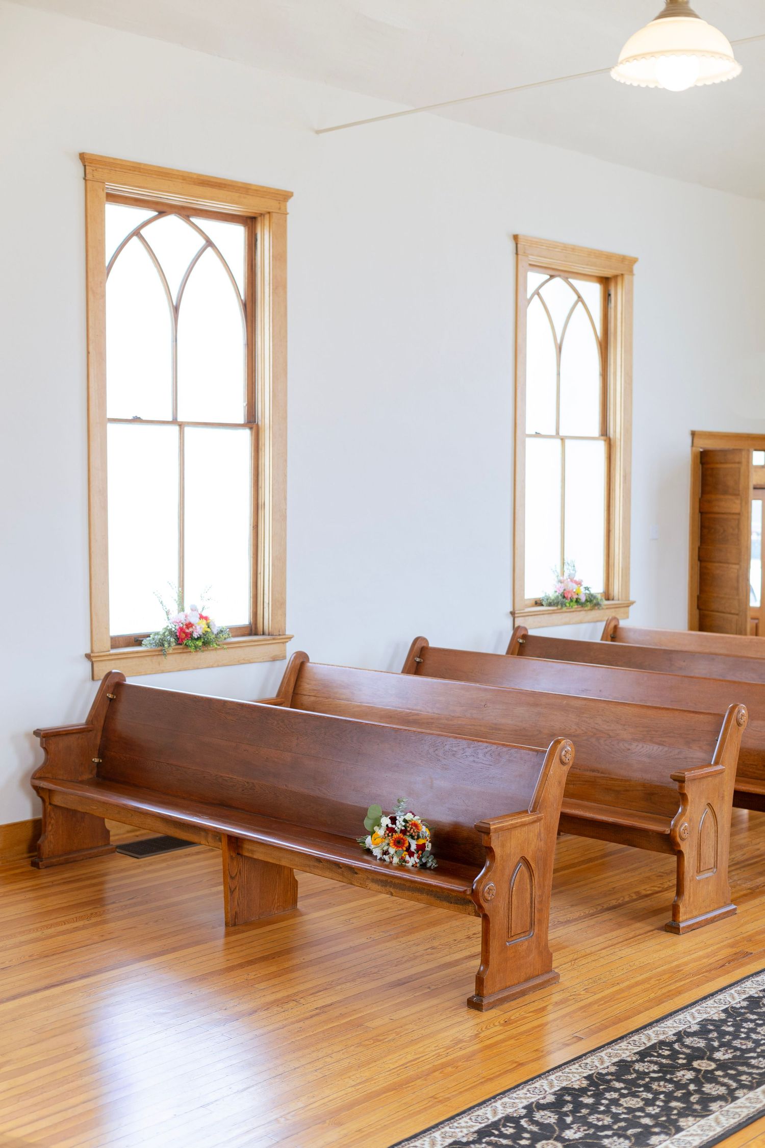 Sunlit church interior with wooden pews, hardwood floors, and arched gothic windows.
