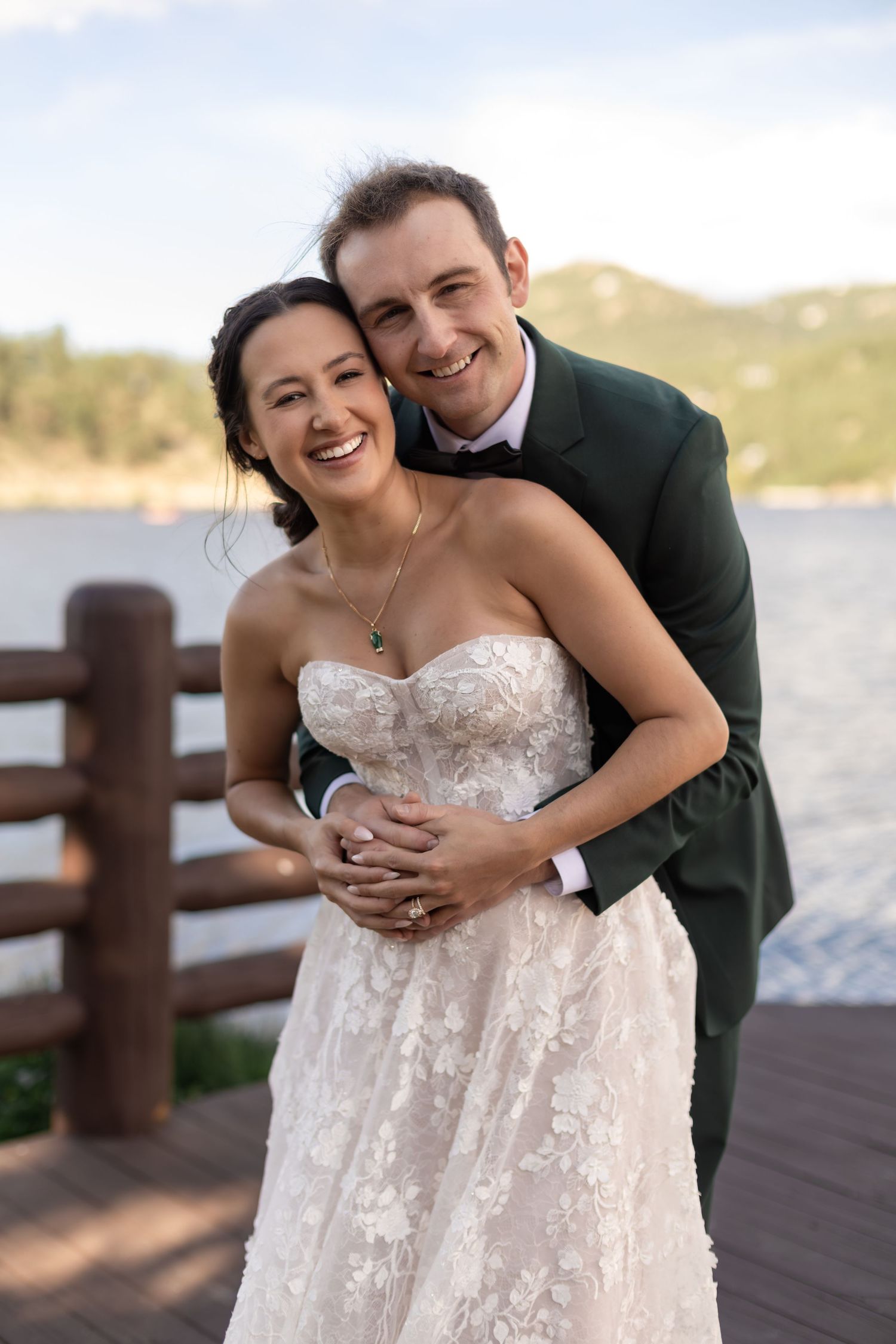 Smiling bride and groom embrace on a lakeside dock, she in a lace gown, he in a green suit.