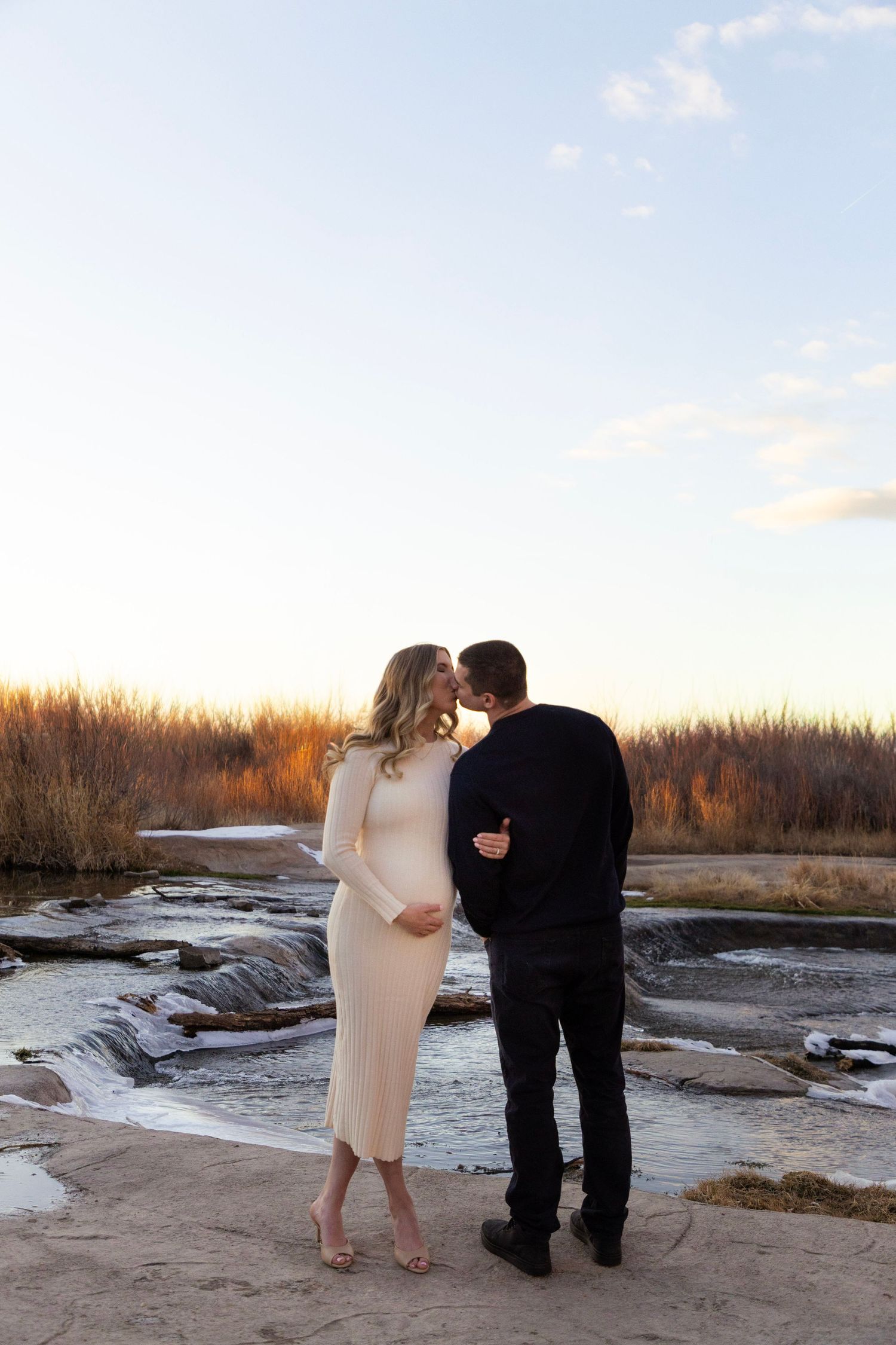 Pregnant couple sharing a kiss outdoors by a rocky stream at sunset, woman in white dress, man in black.