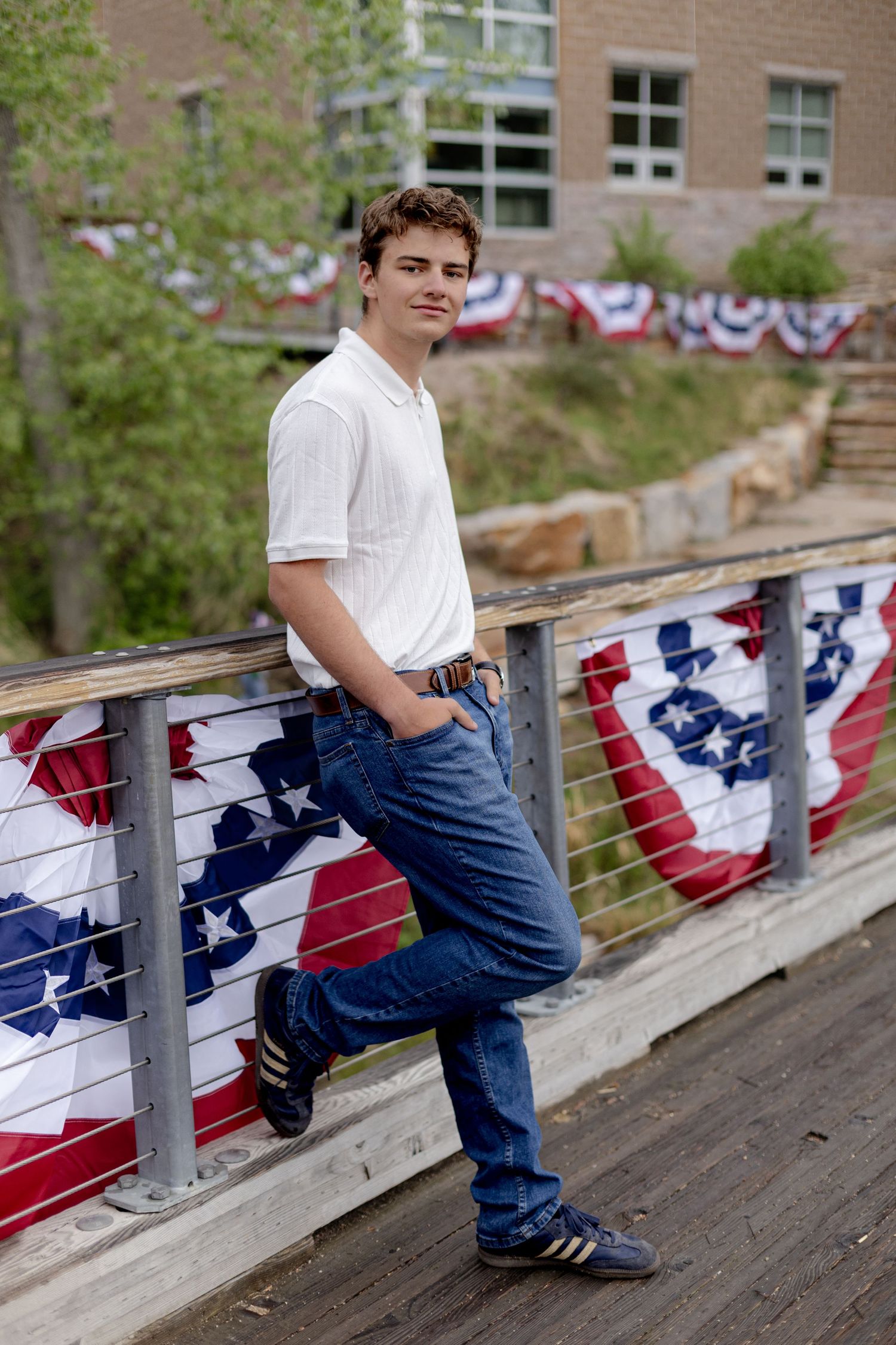 Young man in white polo and jeans leaning on railing decorated with American flags outdoors.