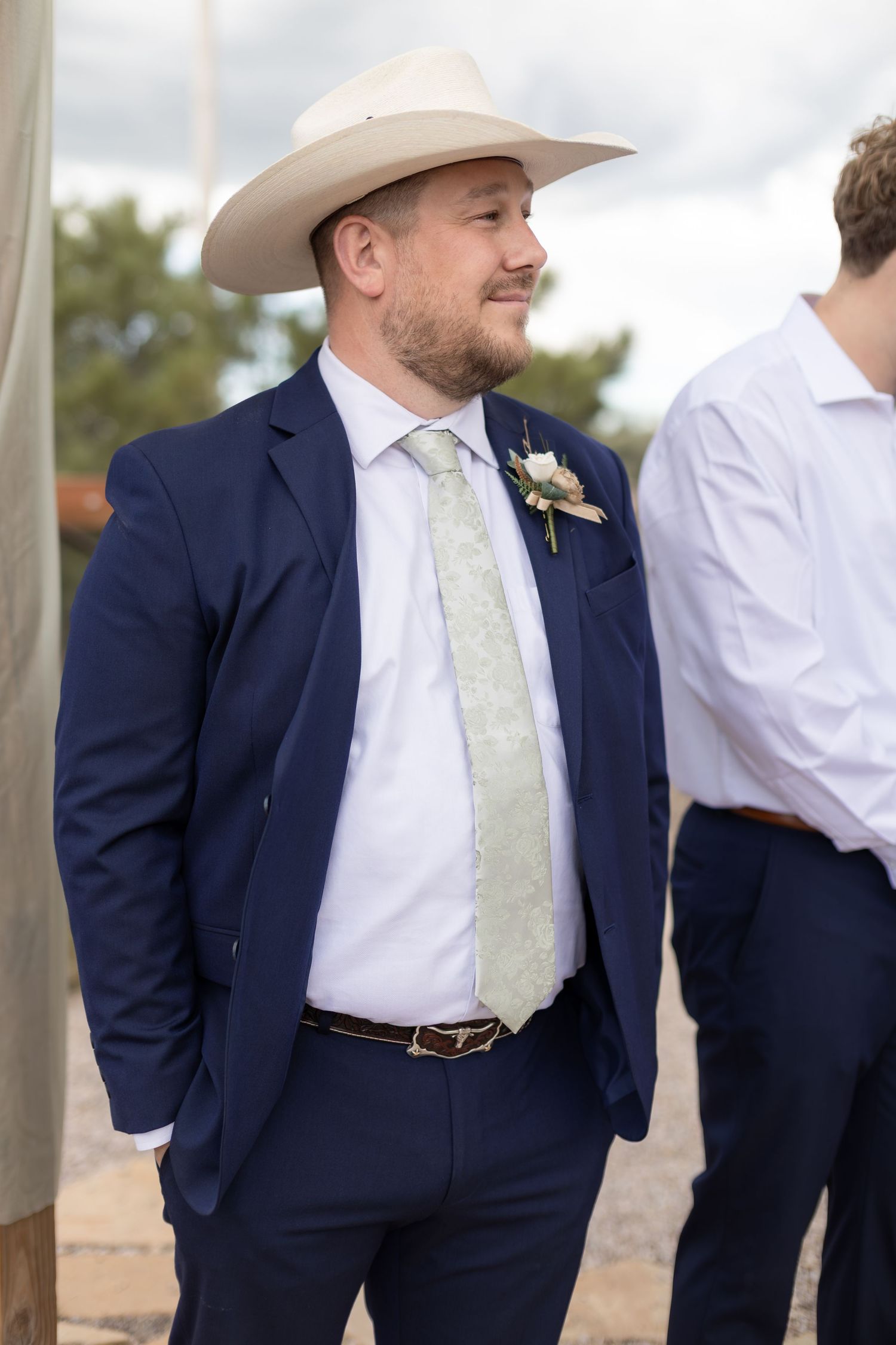 Groom in navy suit, white cowboy hat, floral boutonniere, and sage green tie smiles at outdoor wedding.