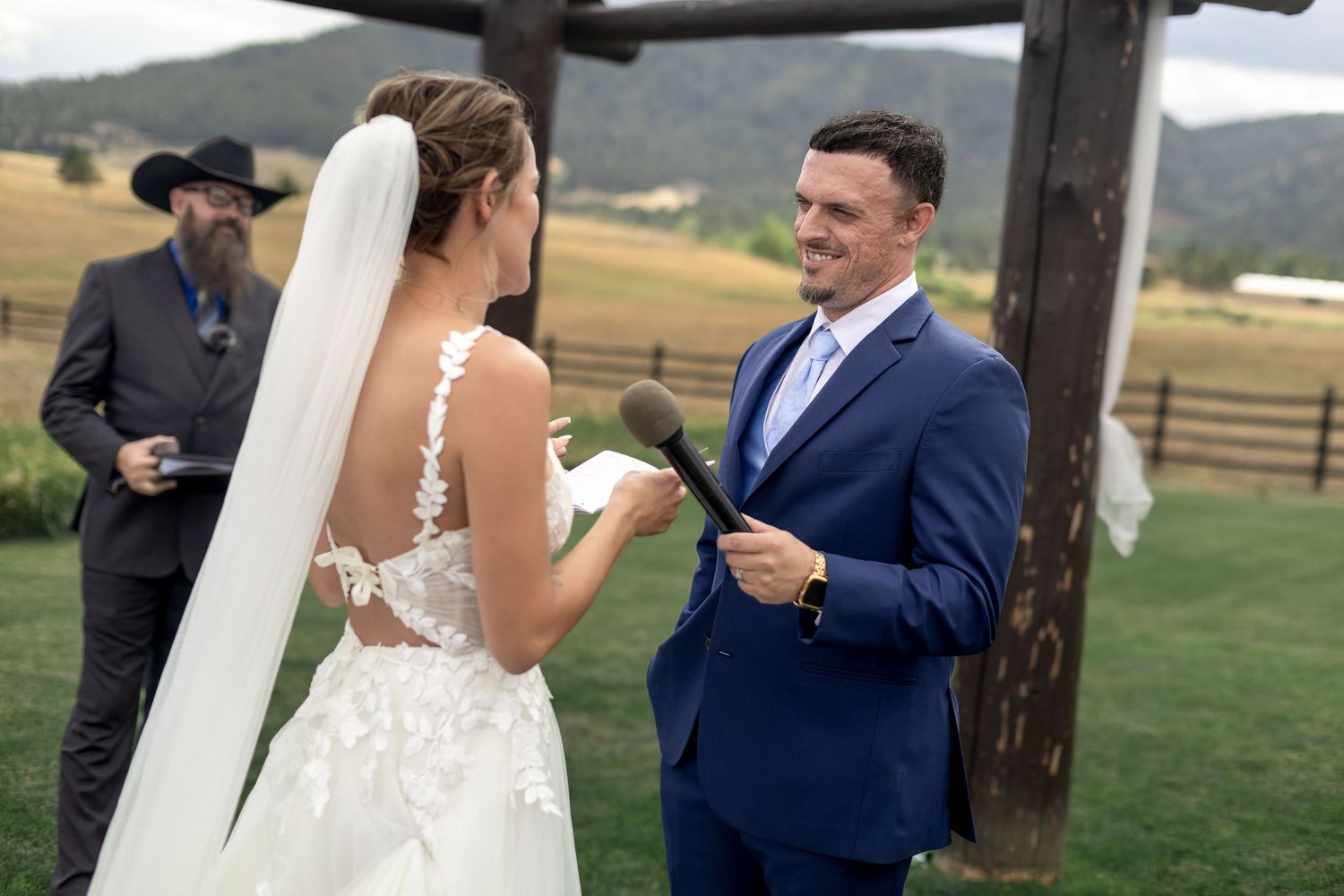 Groom in blue suit reads vows to bride in lace wedding dress at outdoor ranch ceremony with officiant.