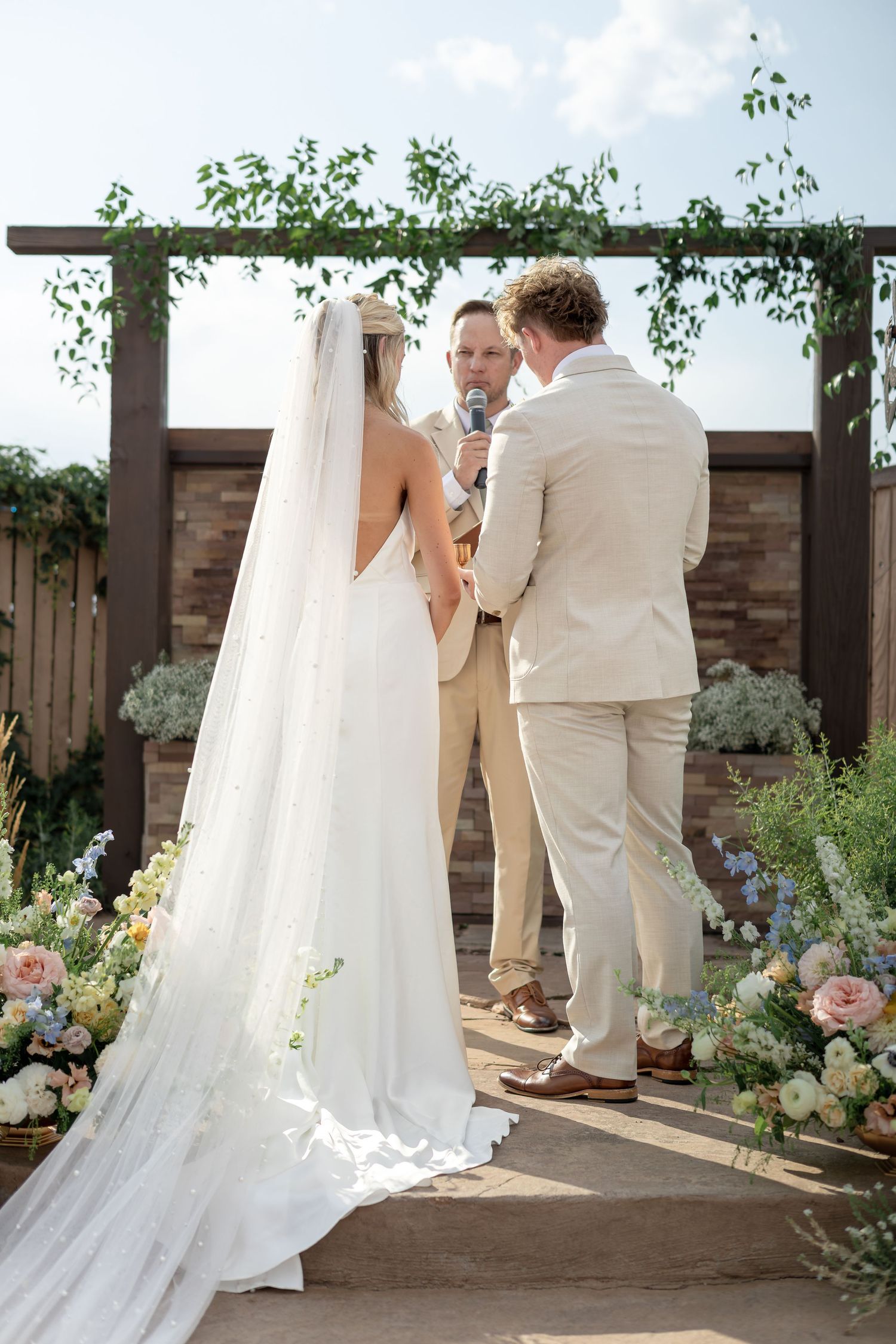 Bride in pearl veil and groom in tan suit exchange vows at floral-adorned outdoor wedding altar.