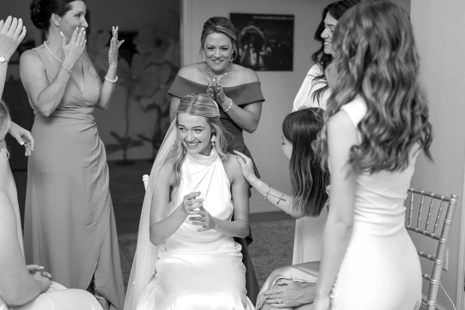 Black and white photo of bride laughing joyfully while bridesmaids help place veil during wedding prep.