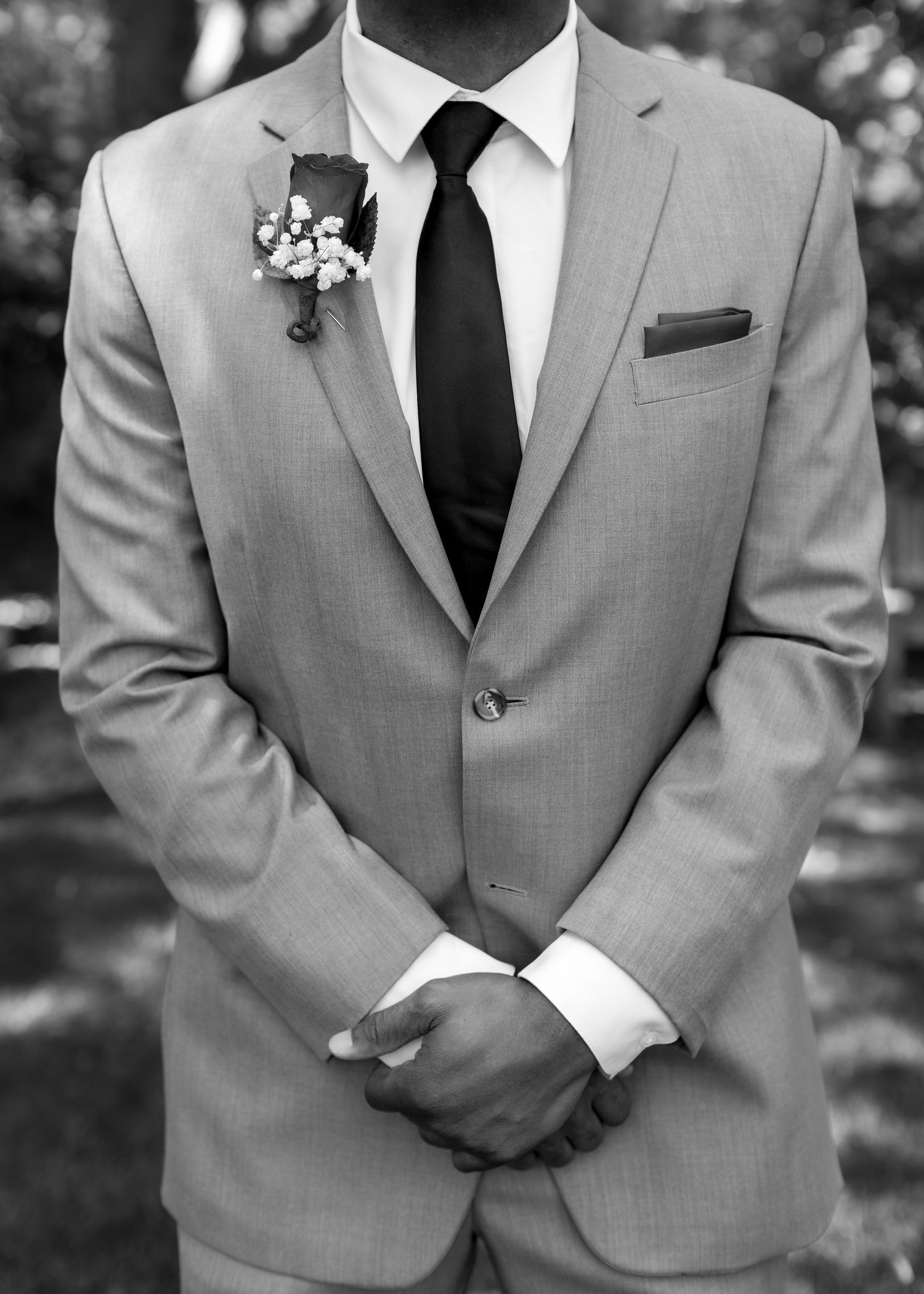 Groom in a light gray suit with black tie and floral boutonniere stands with hands clasped at wedding.