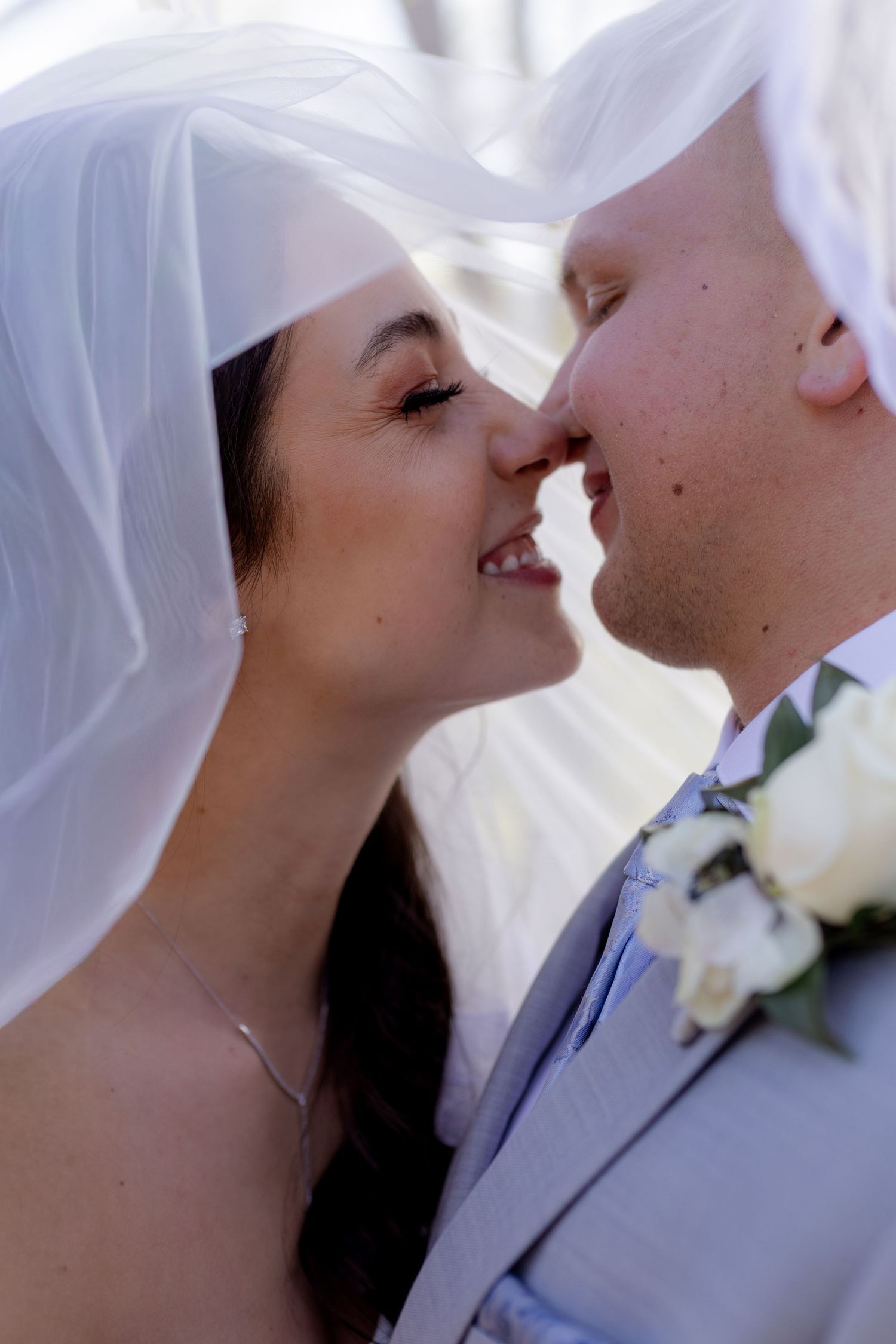 Smiling bride and groom about to kiss under flowing white veil, groom wearing white rose boutonniere.