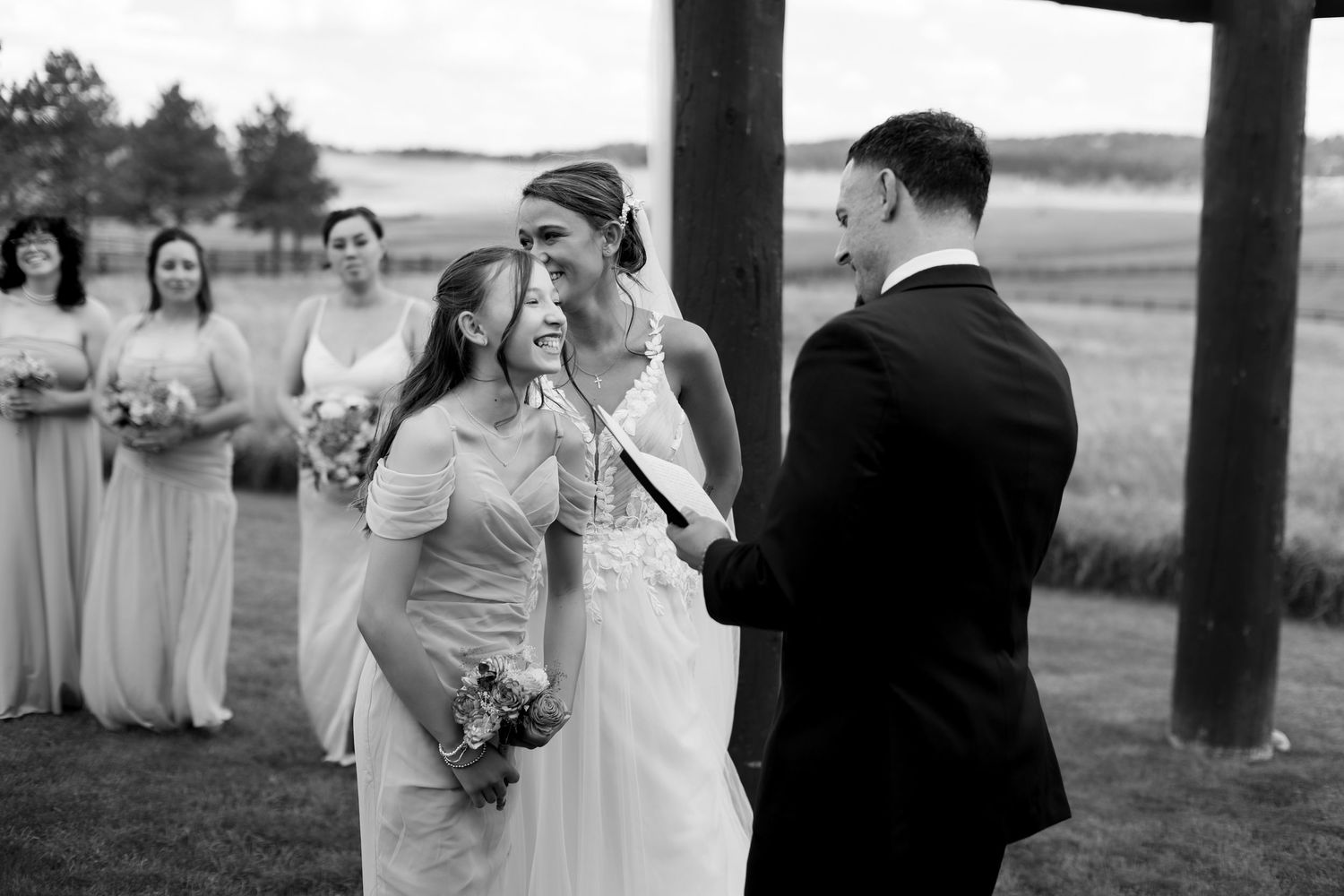 Black and white photo of emotional bride wiping tears during outdoor wedding ceremony with bridesmaids standing behind.