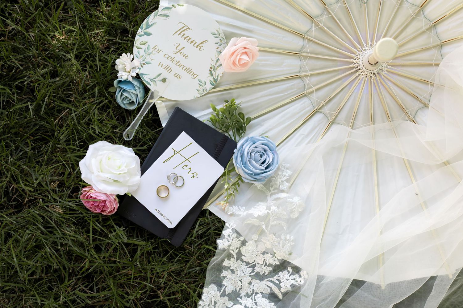 Wedding flat lay on grass featuring a parasol, floral boutonnieres, lace veil, earrings, and a decorative fan.