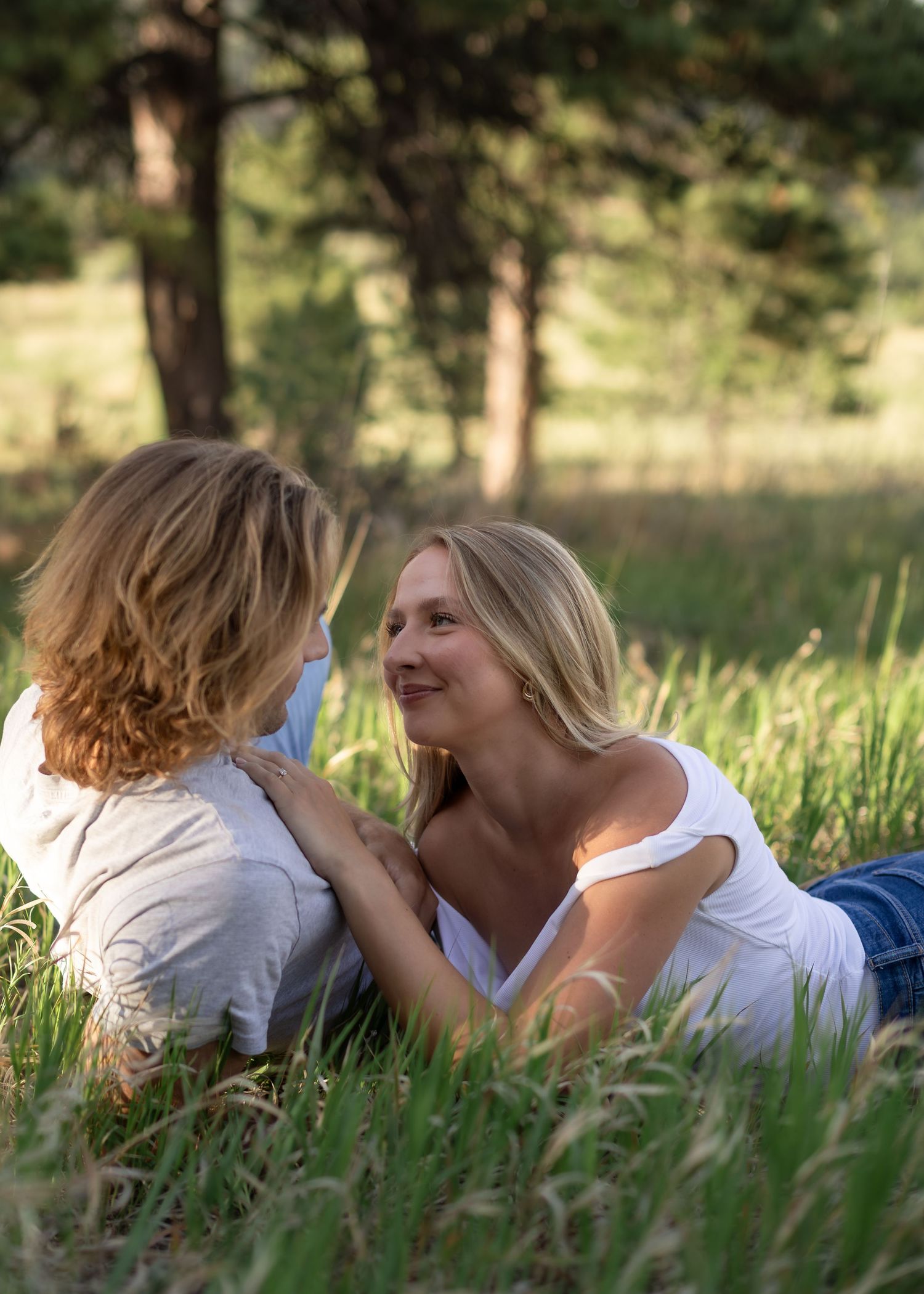 A mother and child share a tender moment lying in green grass with pine trees in the background.