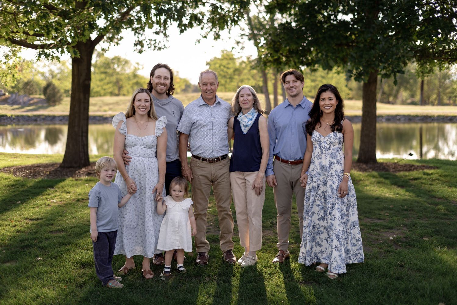 A multigenerational family of seven poses together outdoors near a pond surrounded by lush green trees.