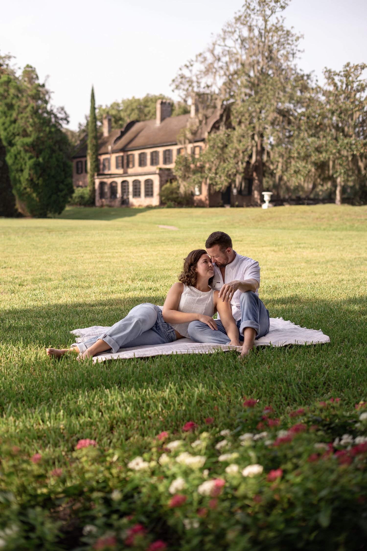 Romantic couple sits together on blanket in lush garden with historic brick mansion in background.