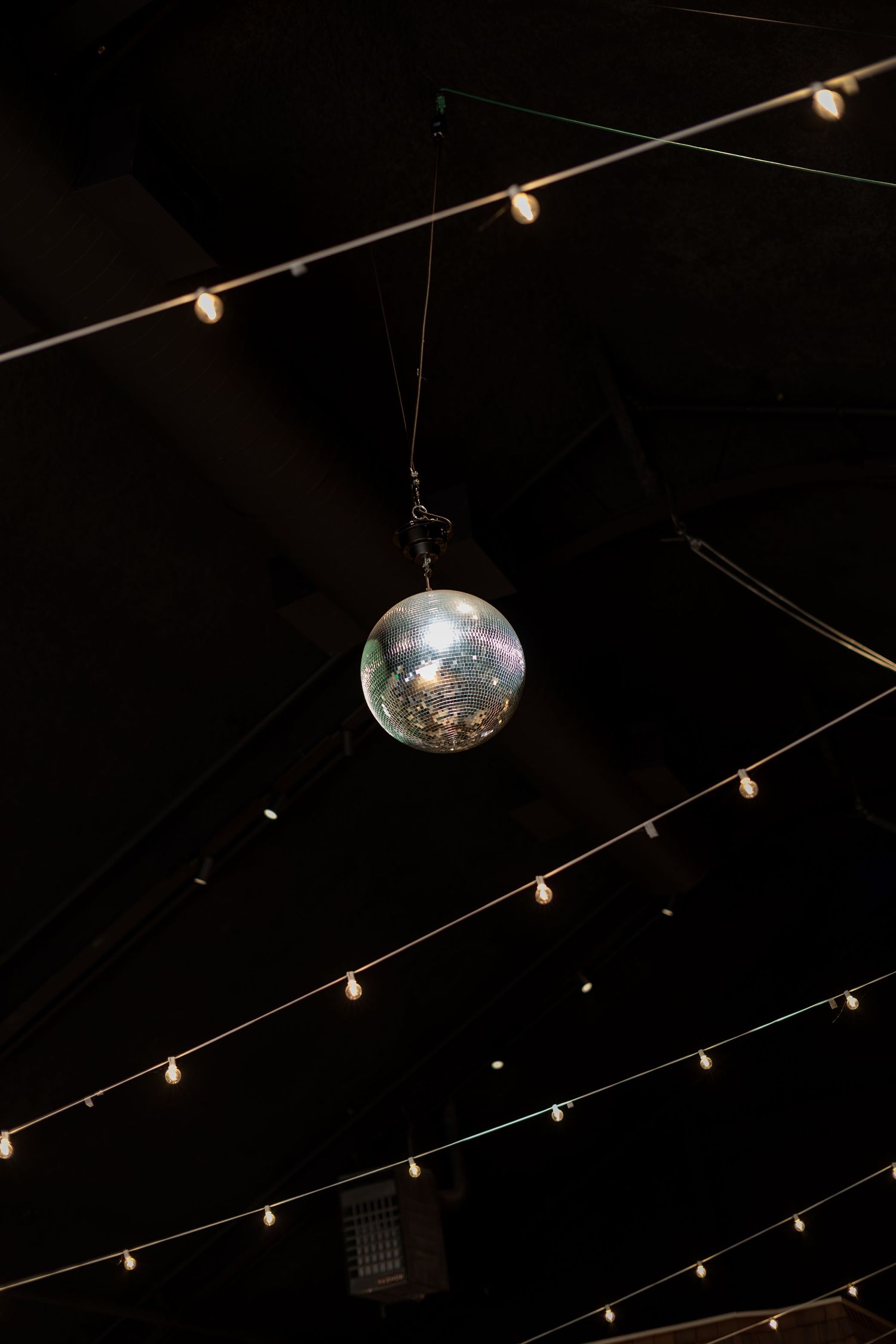A shimmering disco ball hangs from a dark ceiling surrounded by warm string lights.