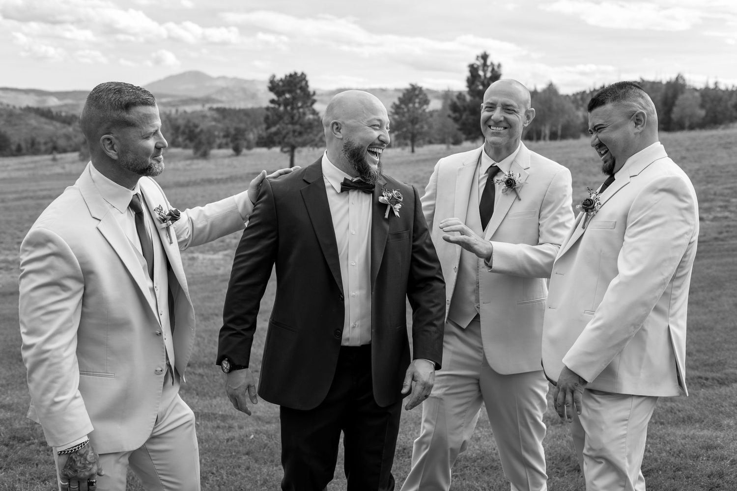 Black and white photo of groom in dark tuxedo laughing with three groomsmen in light suits outdoors.
