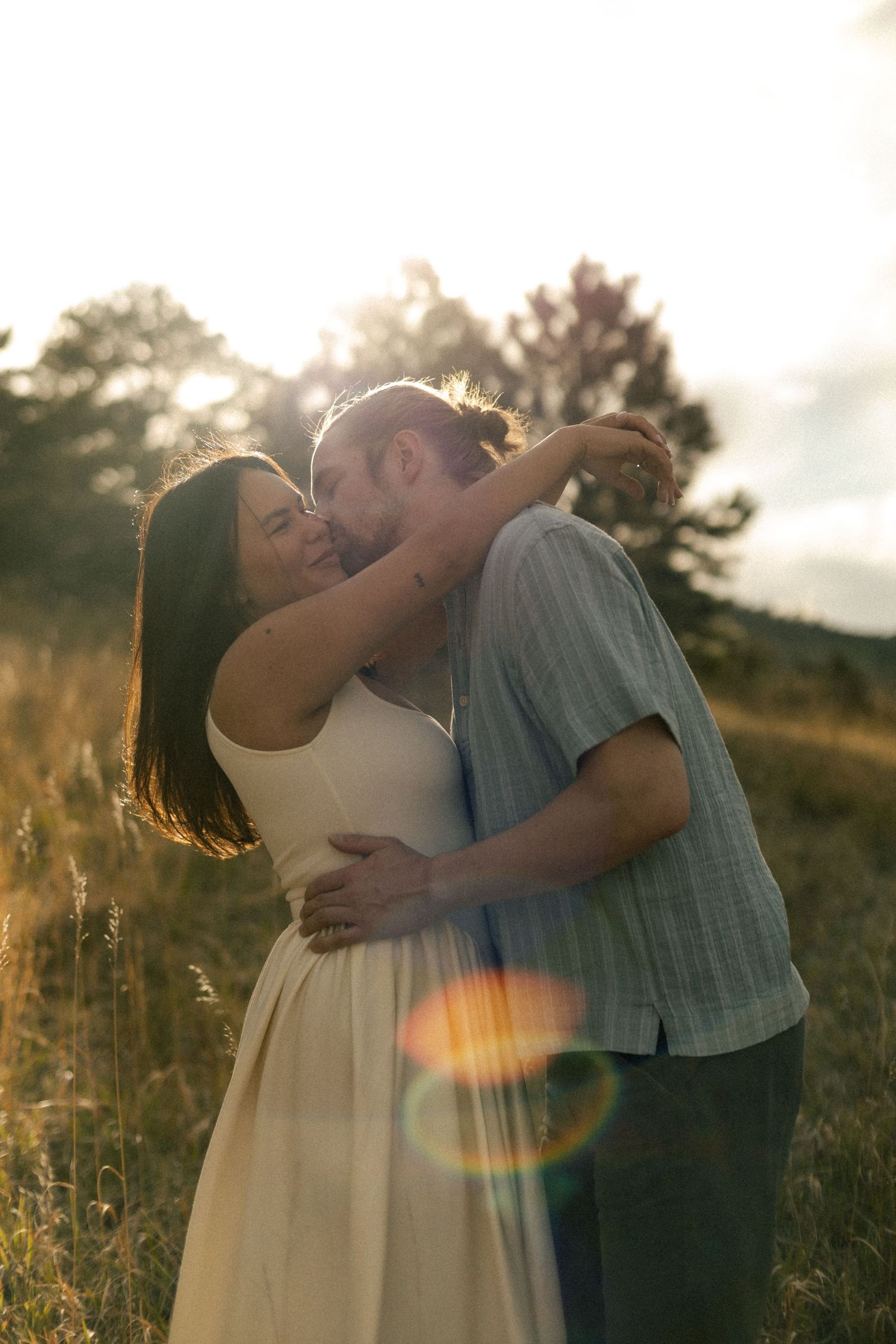Couple embracing and kissing in a sunlit meadow, golden hour lens flare, woman in white dress.