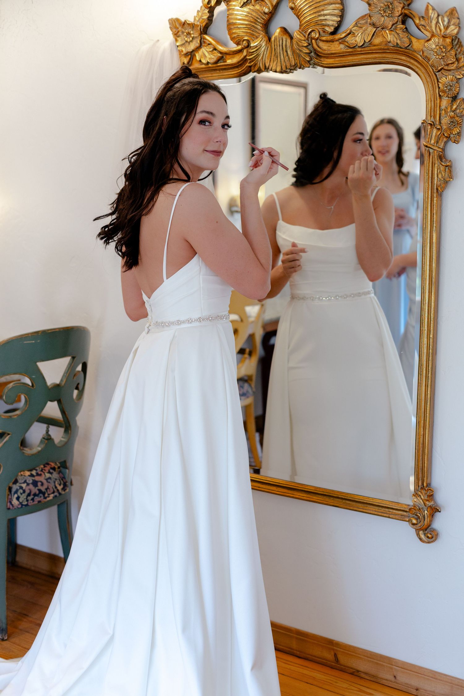 Bride in white satin gown with spaghetti straps applies makeup while looking in ornate gold mirror.