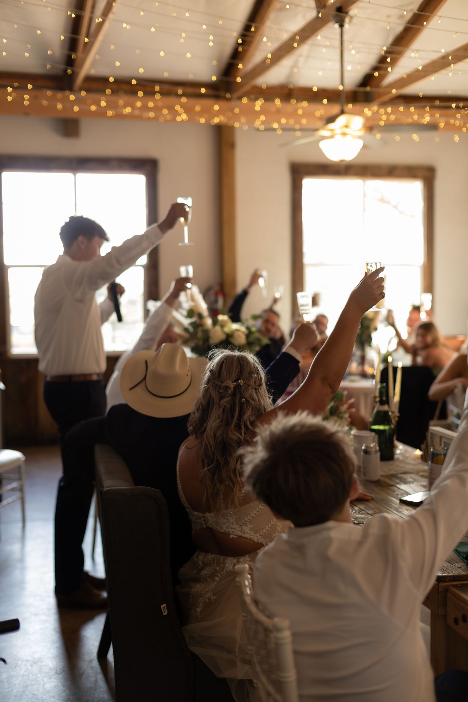 Wedding guests raise glasses in toast inside rustic venue decorated with string lights during reception.