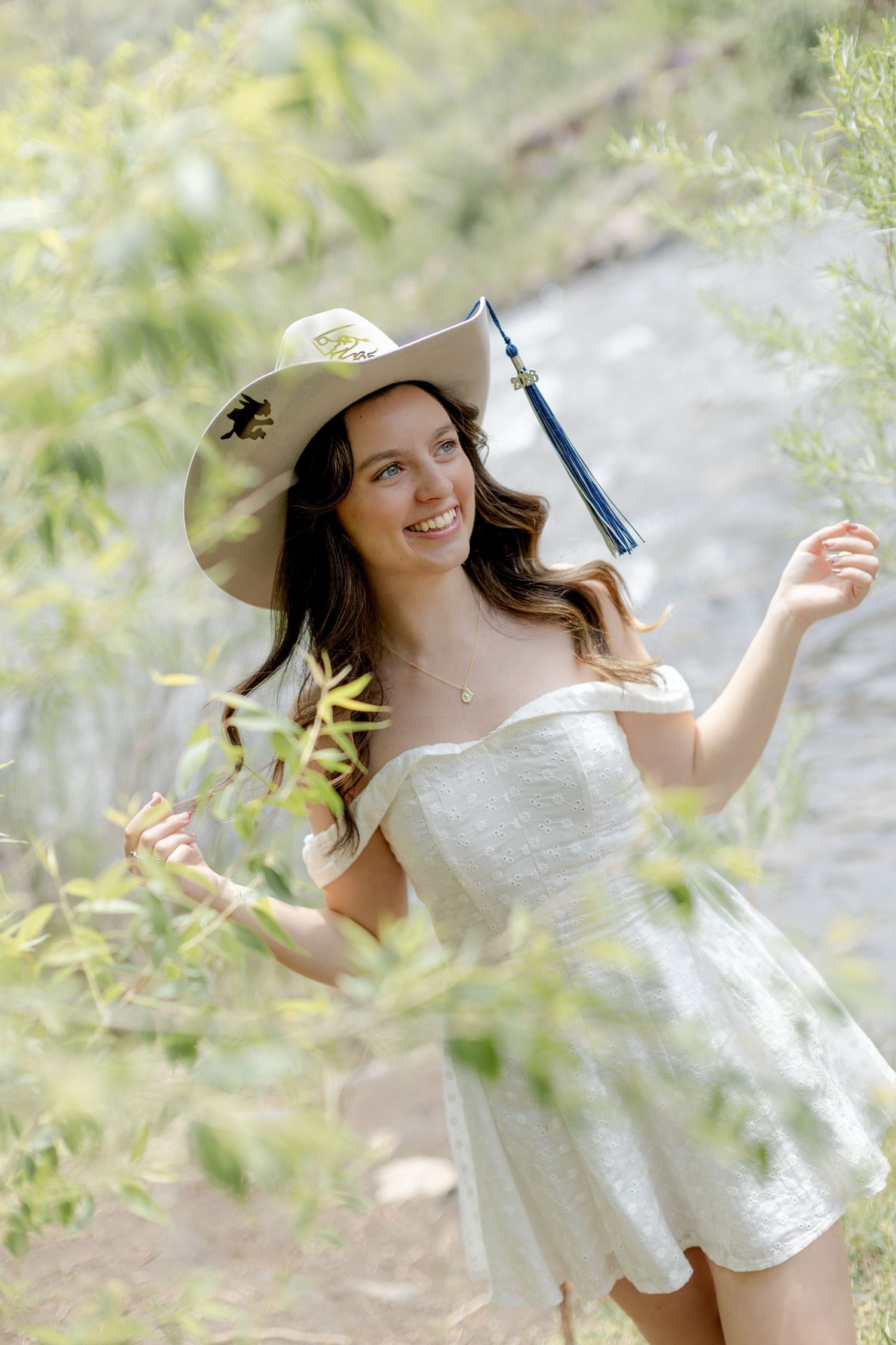 Smiling young woman in white off-shoulder dress and graduation cap with tassel poses outdoors among green foliage.