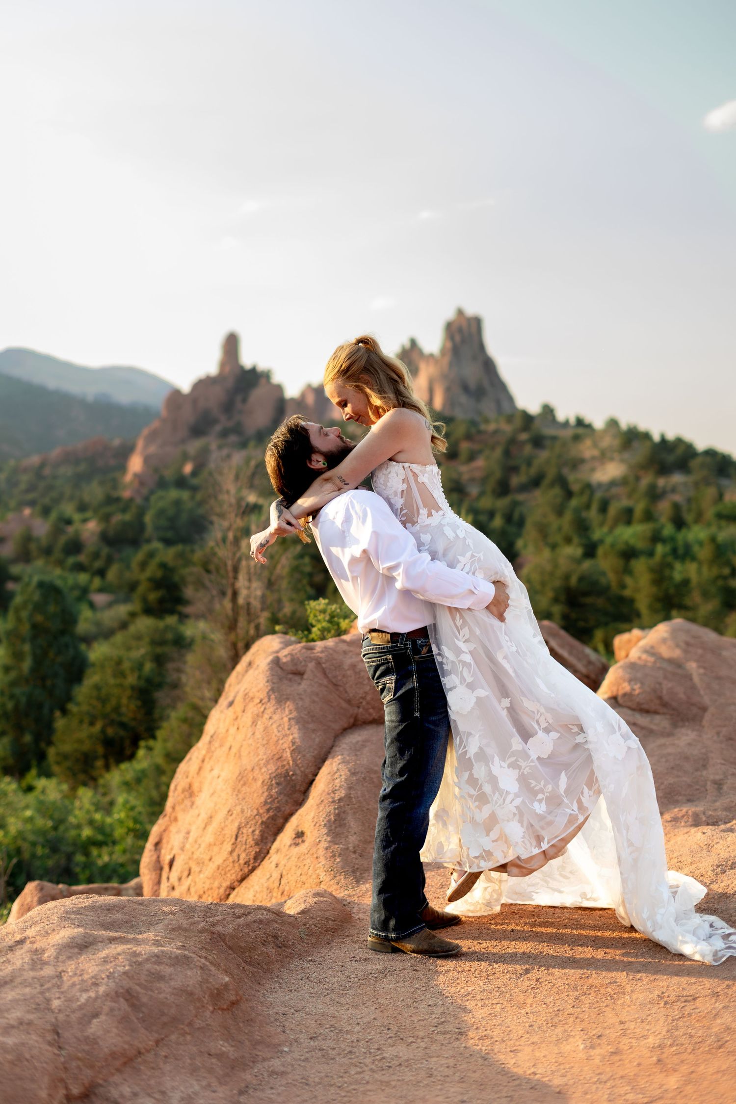 Groom lifts bride in floral wedding dress at Garden of the Gods, Colorado, with red rock formations behind them.
