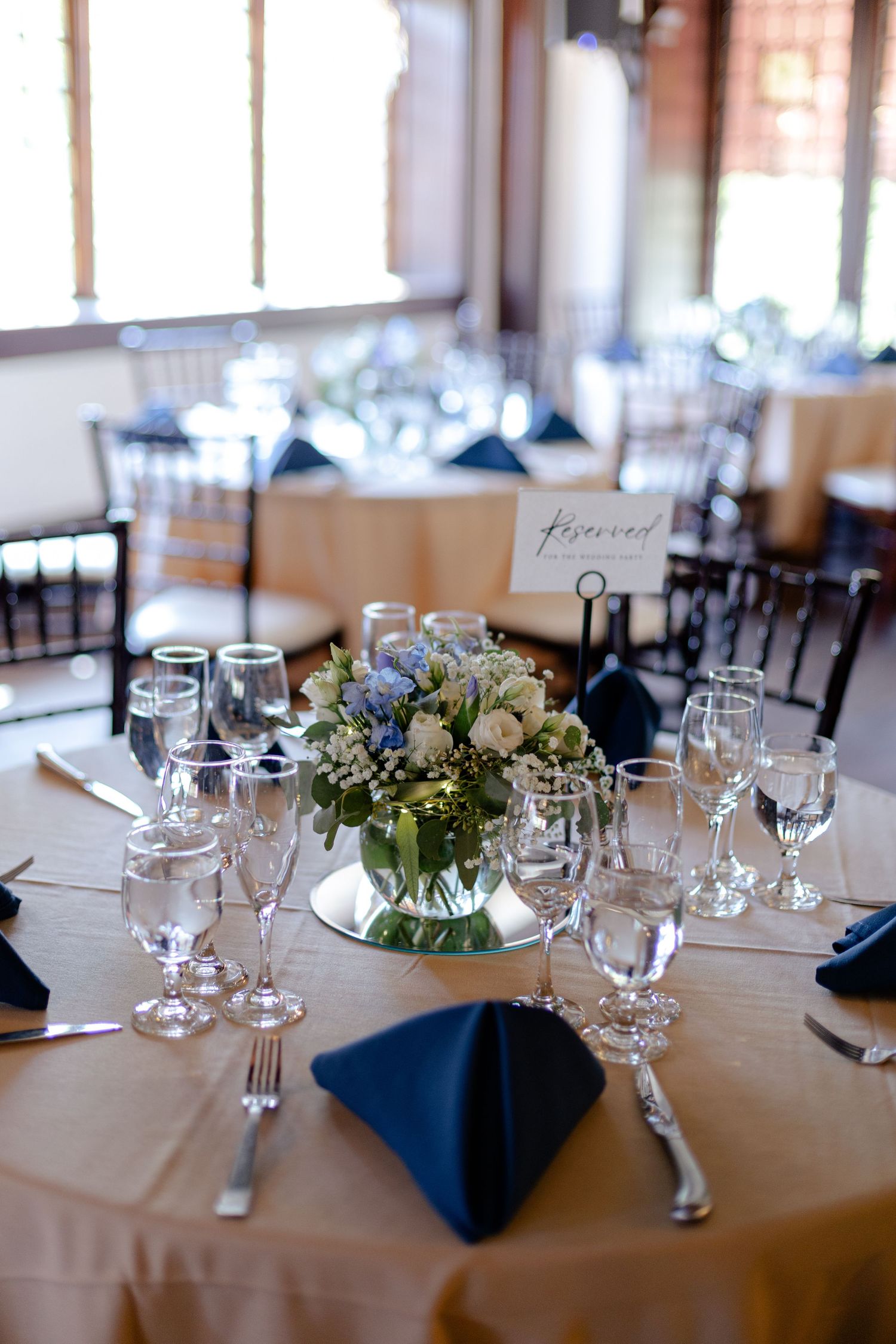 Wedding reception table set with champagne linens, navy napkins, and a centerpiece of white and blue flowers on a mirror.