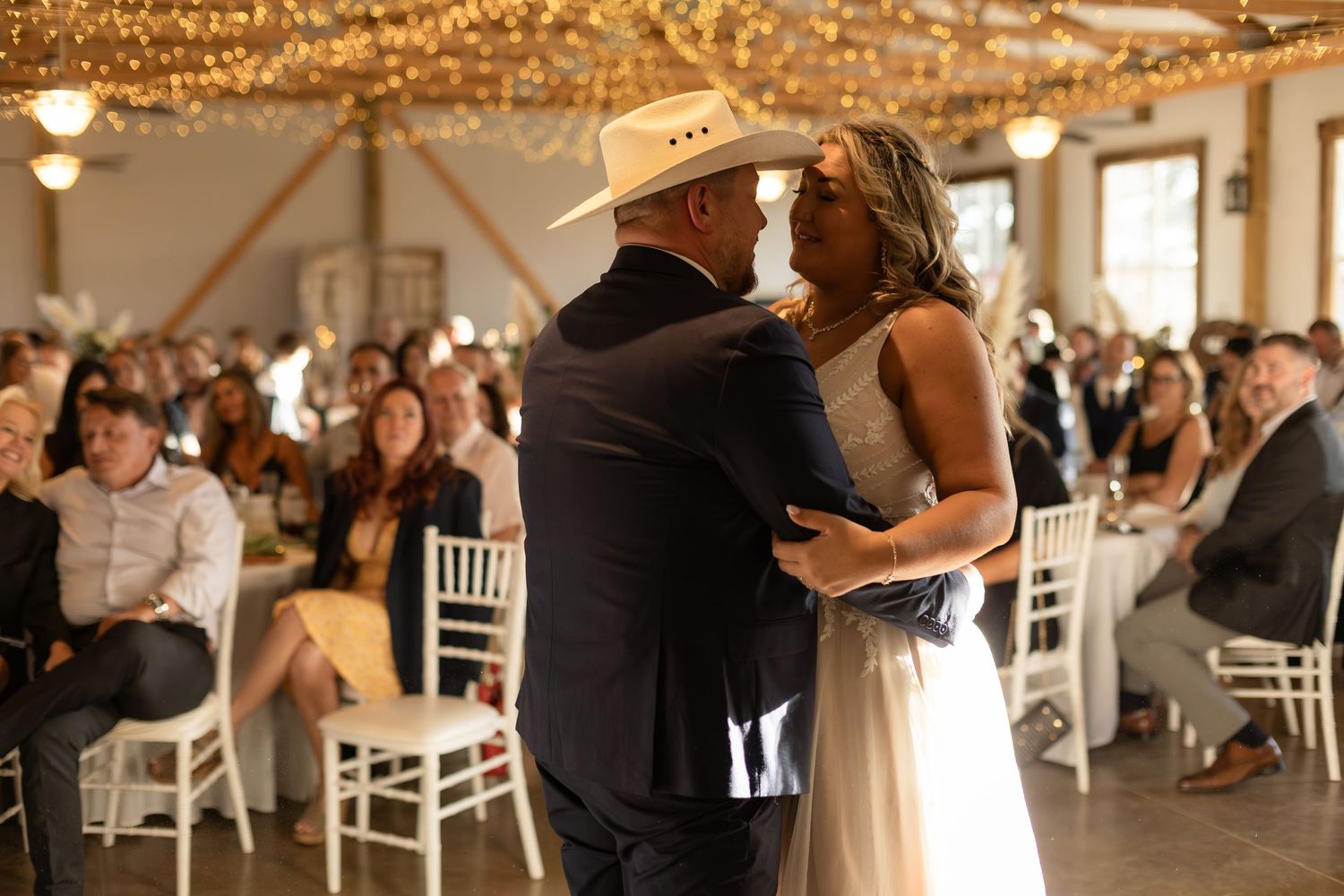 Bride and groom in cowboy hat share first dance before seated wedding guests in decorated barn venue.