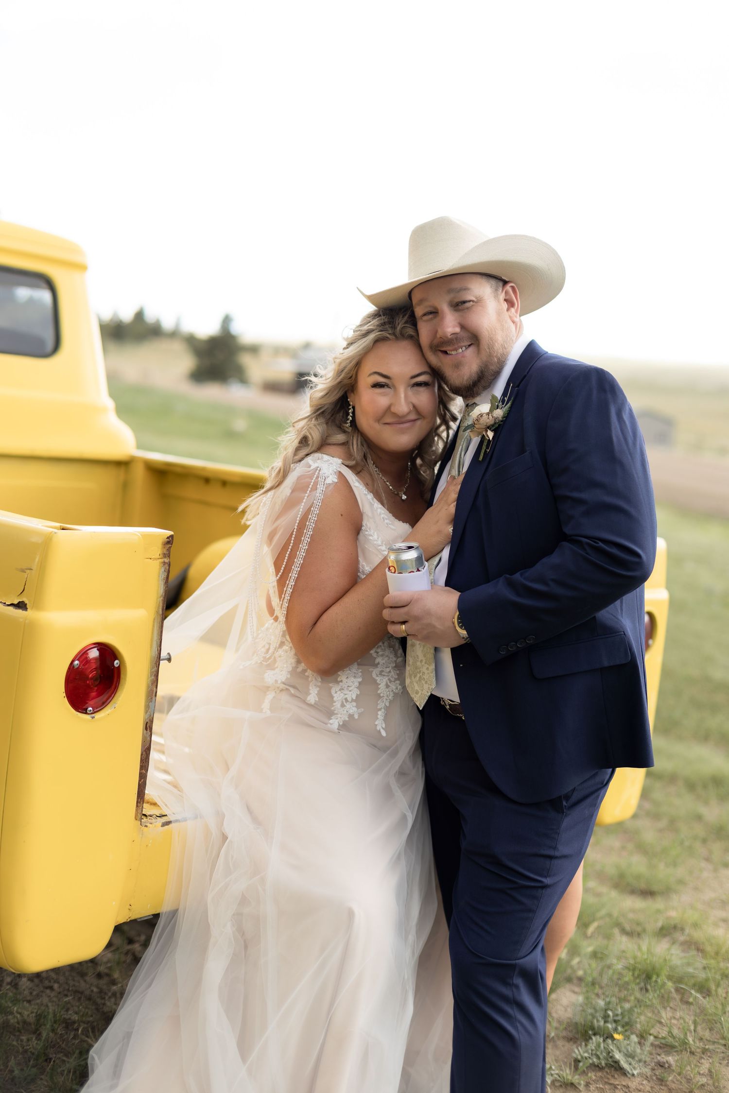Bride and groom posing together beside a vintage yellow pickup truck at an outdoor country wedding.