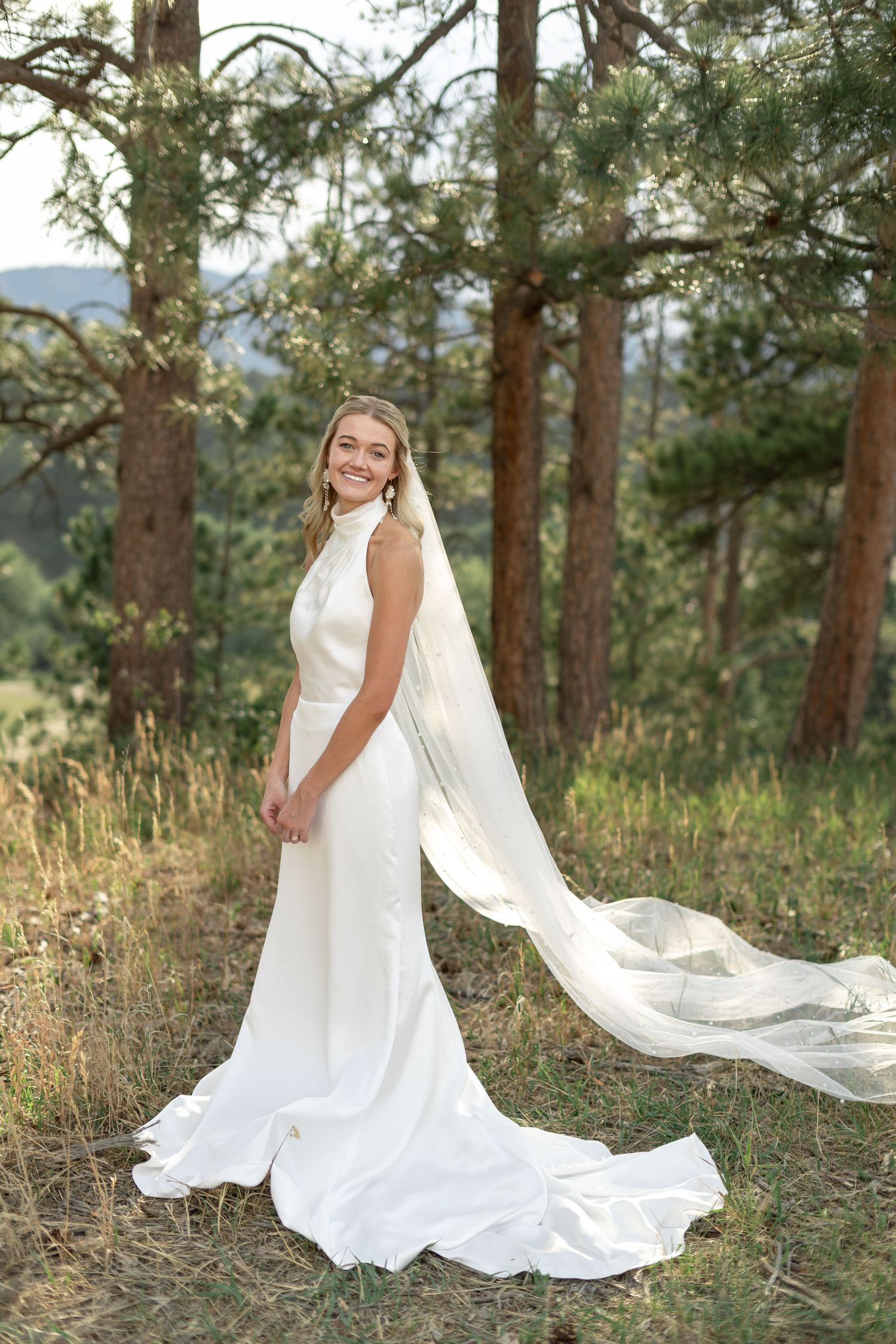 Elegant bride in sleek white halter gown with long flowing veil poses among pine trees outdoors.