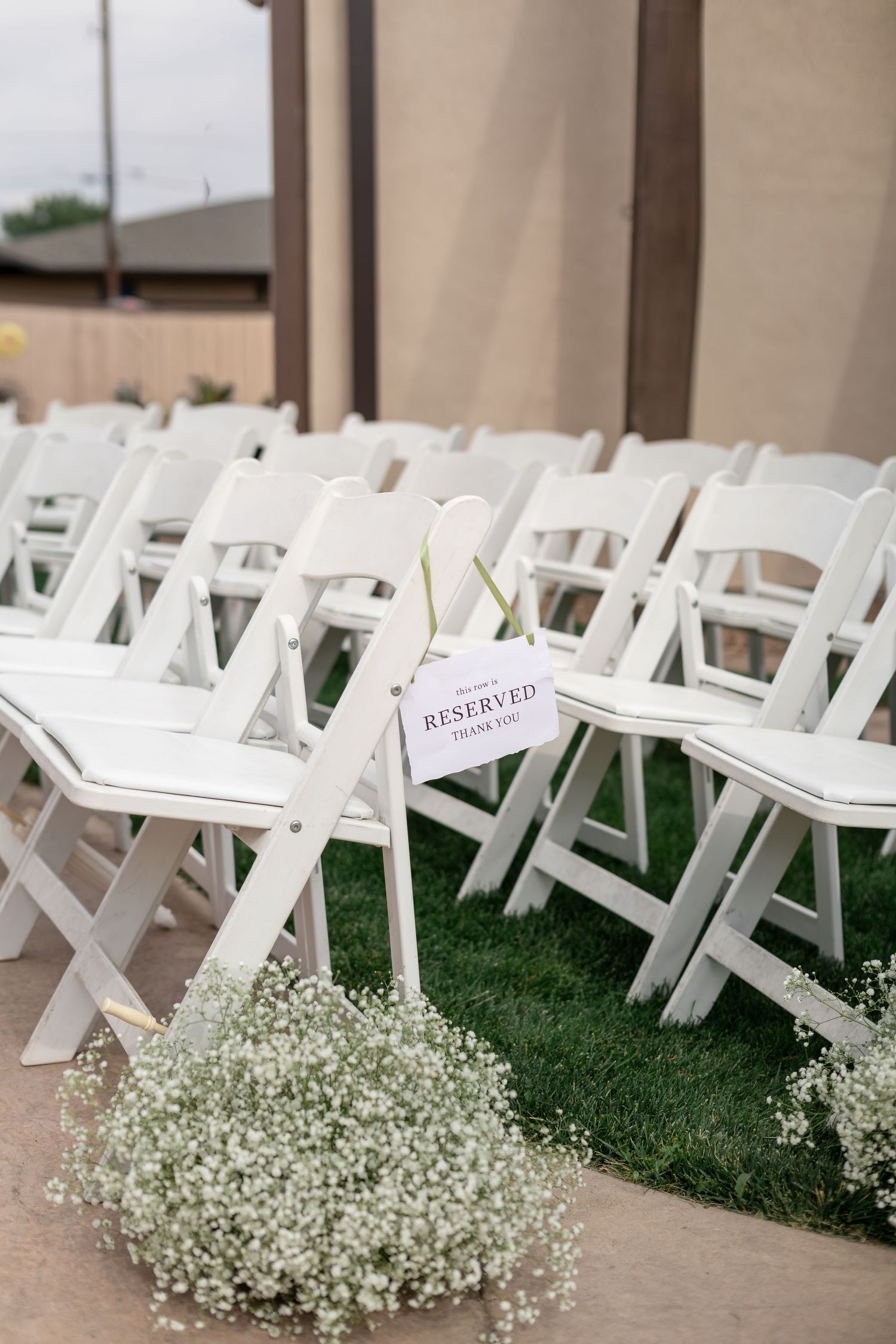Rows of white folding chairs with reserved sign and baby's breath floral arrangements at outdoor wedding ceremony.