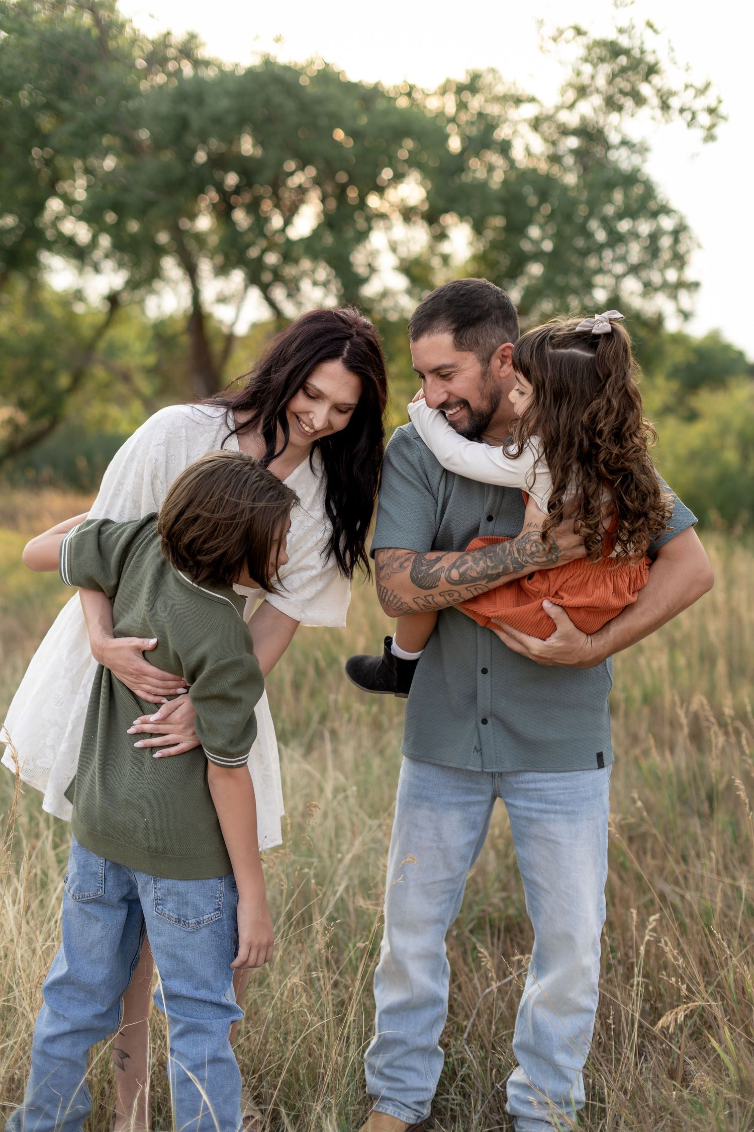 Happy family of four sharing a joyful moment in a grassy field, parents embracing their two children outdoors.