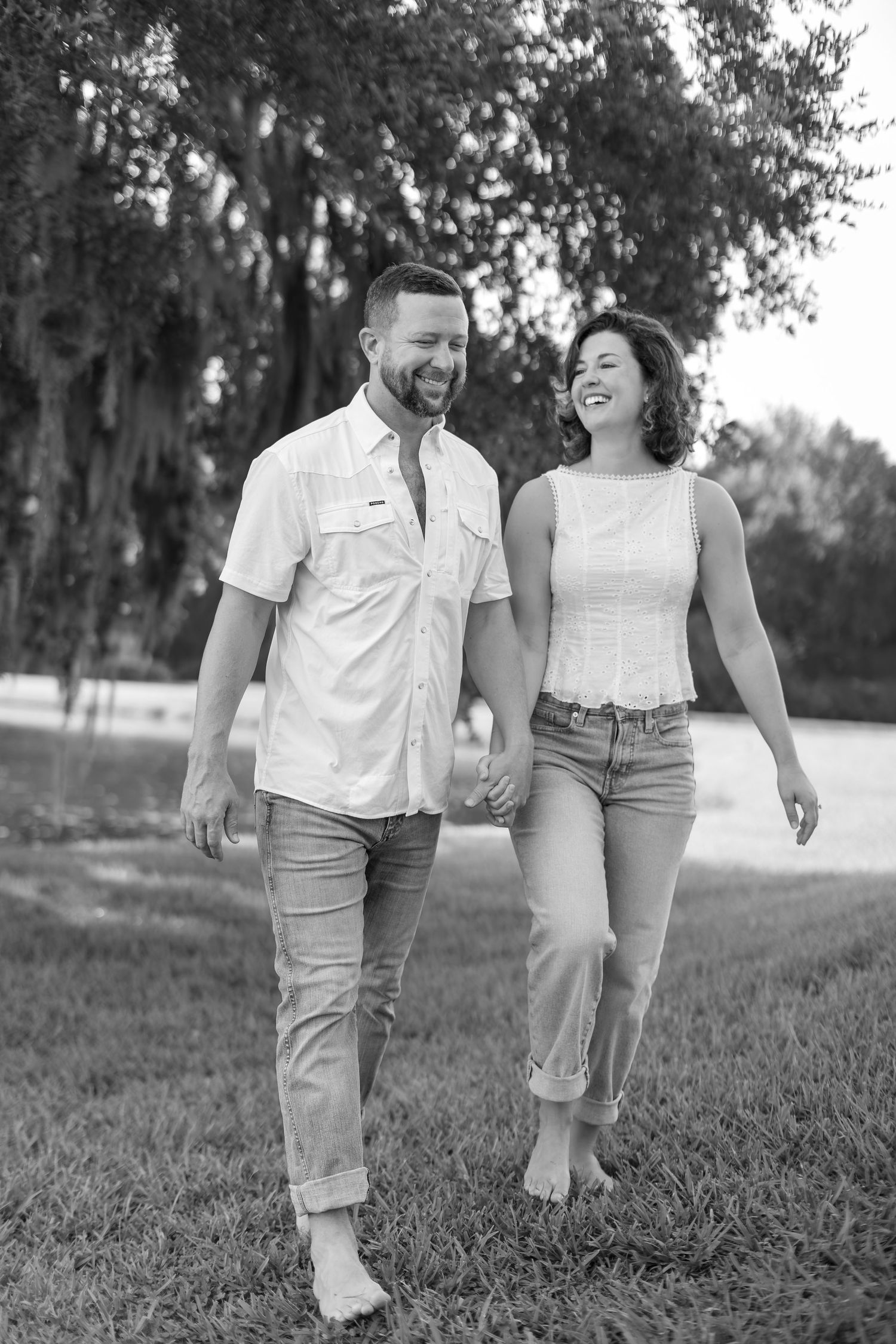 Black and white photo of a smiling couple holding hands and walking barefoot on grass with trees in the background.