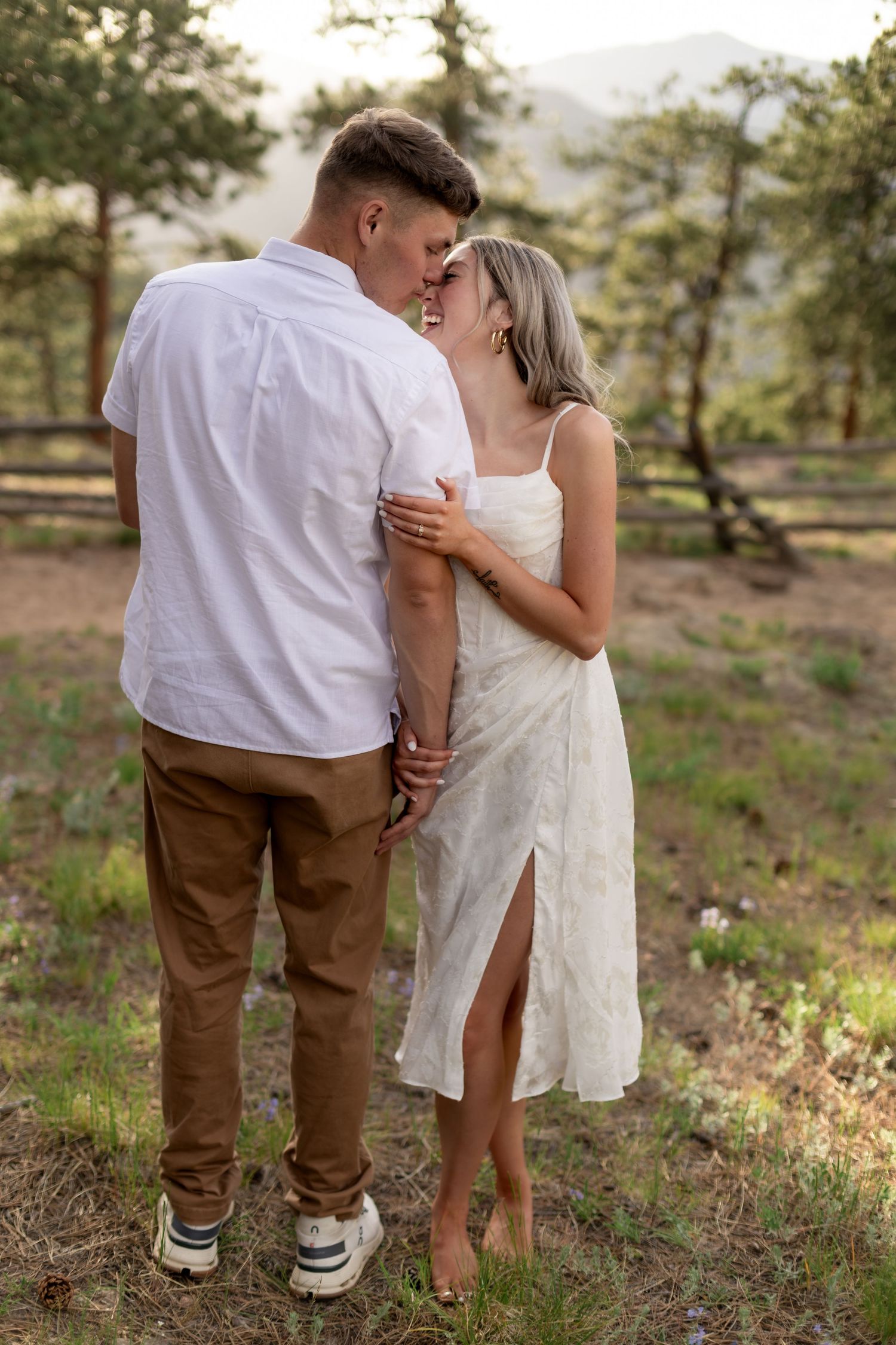 Couple sharing a romantic moment outdoors, woman in white dress and man in white shirt, surrounded by trees and mountains.