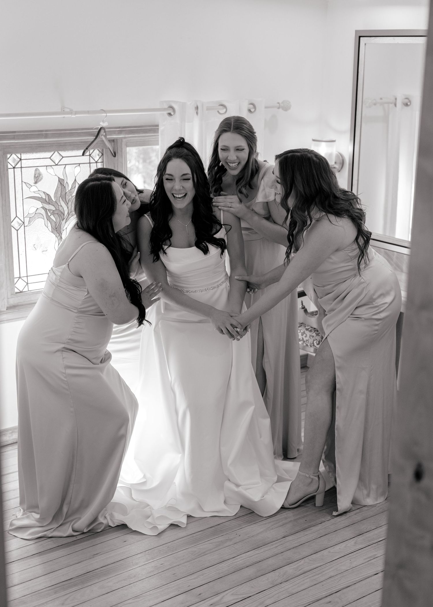 Bride in white gown laughing with three bridesmaids in light dresses during wedding preparations indoors.