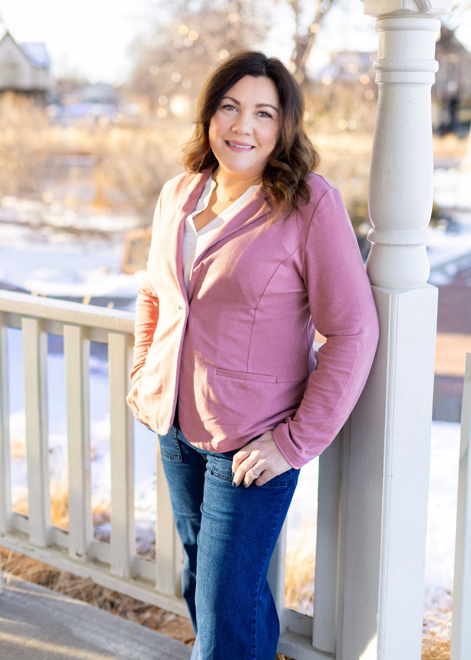 Woman in pink blazer and jeans smiling outdoors on a porch with snowy background.