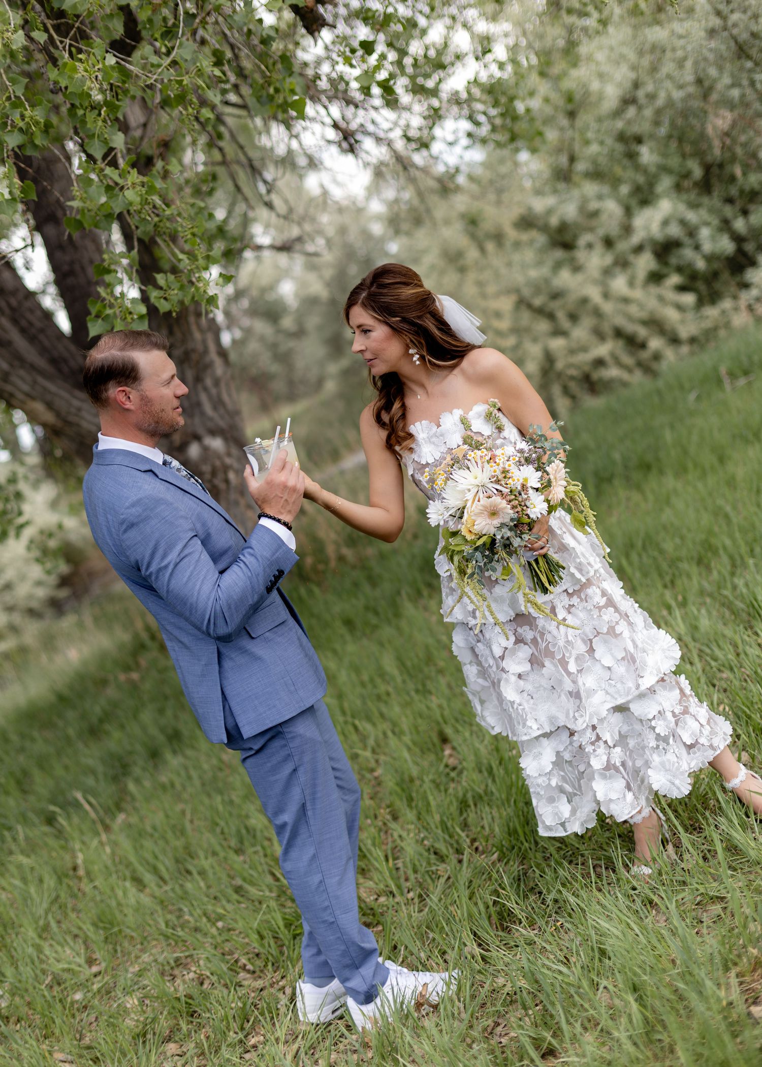 Groom in blue suit toasts with bride holding bouquet in lush green outdoor wedding setting.