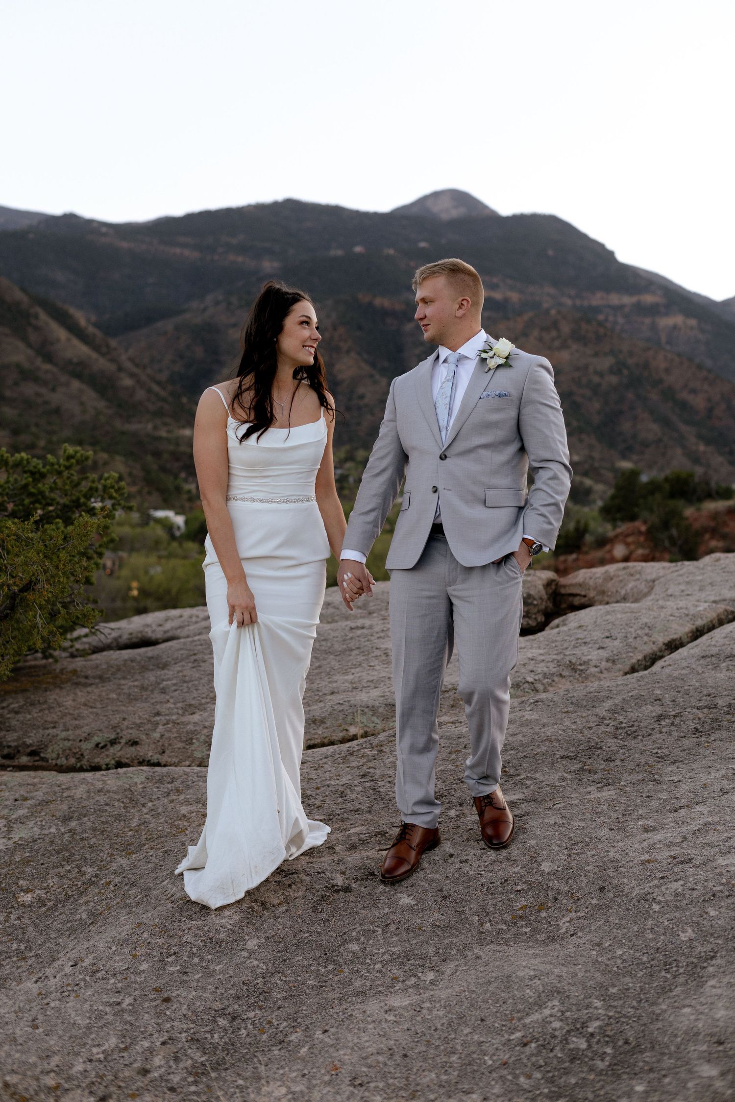 Bride and groom holding hands on rocky mountain overlook with scenic mountain range backdrop.