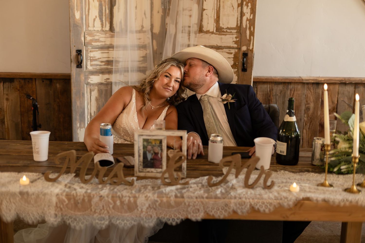 Bride and groom in cowboy hat share intimate moment at rustic sweetheart table with candles and decor.