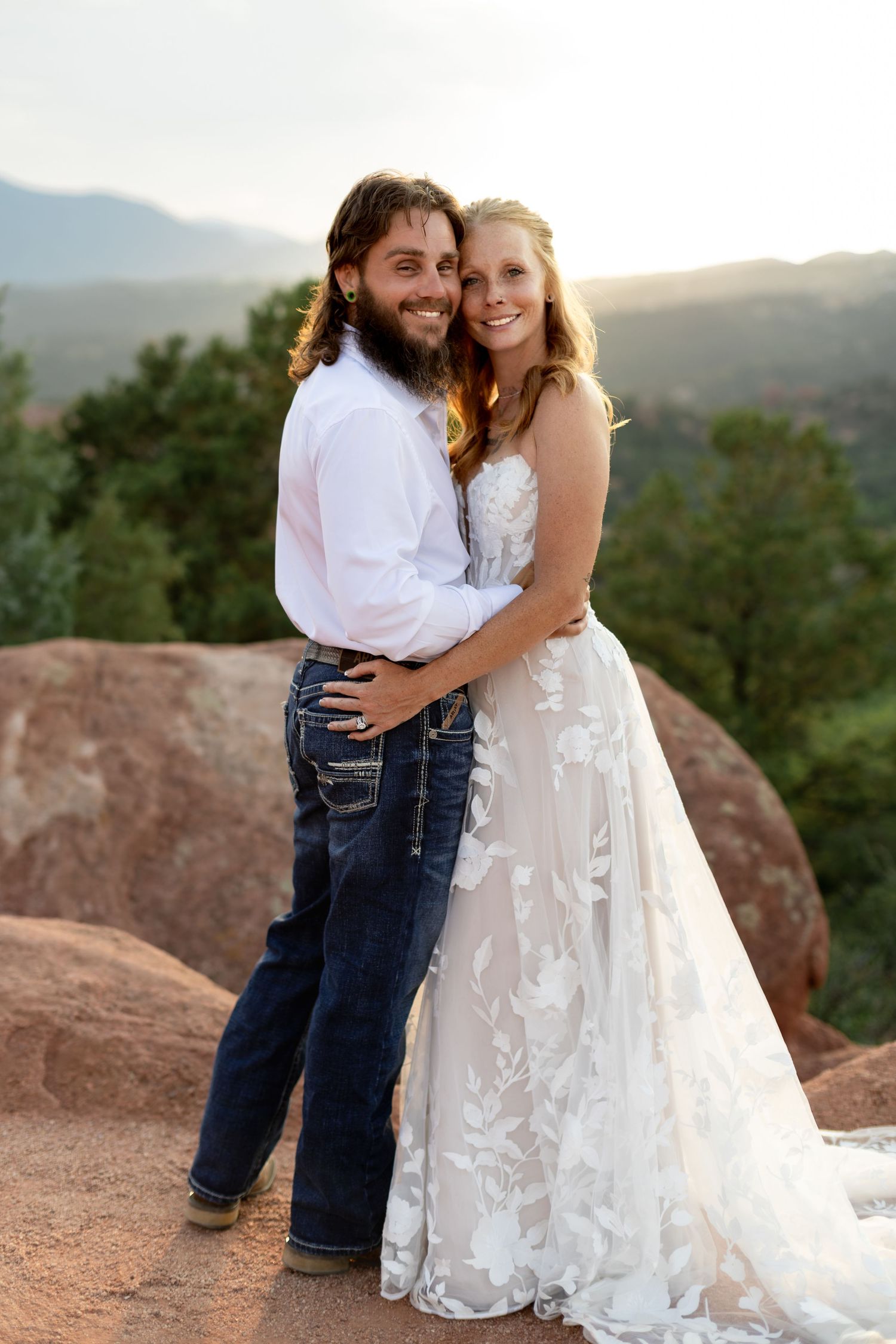 Smiling bride and groom pose together in wedding attire on red rocks at golden hour.