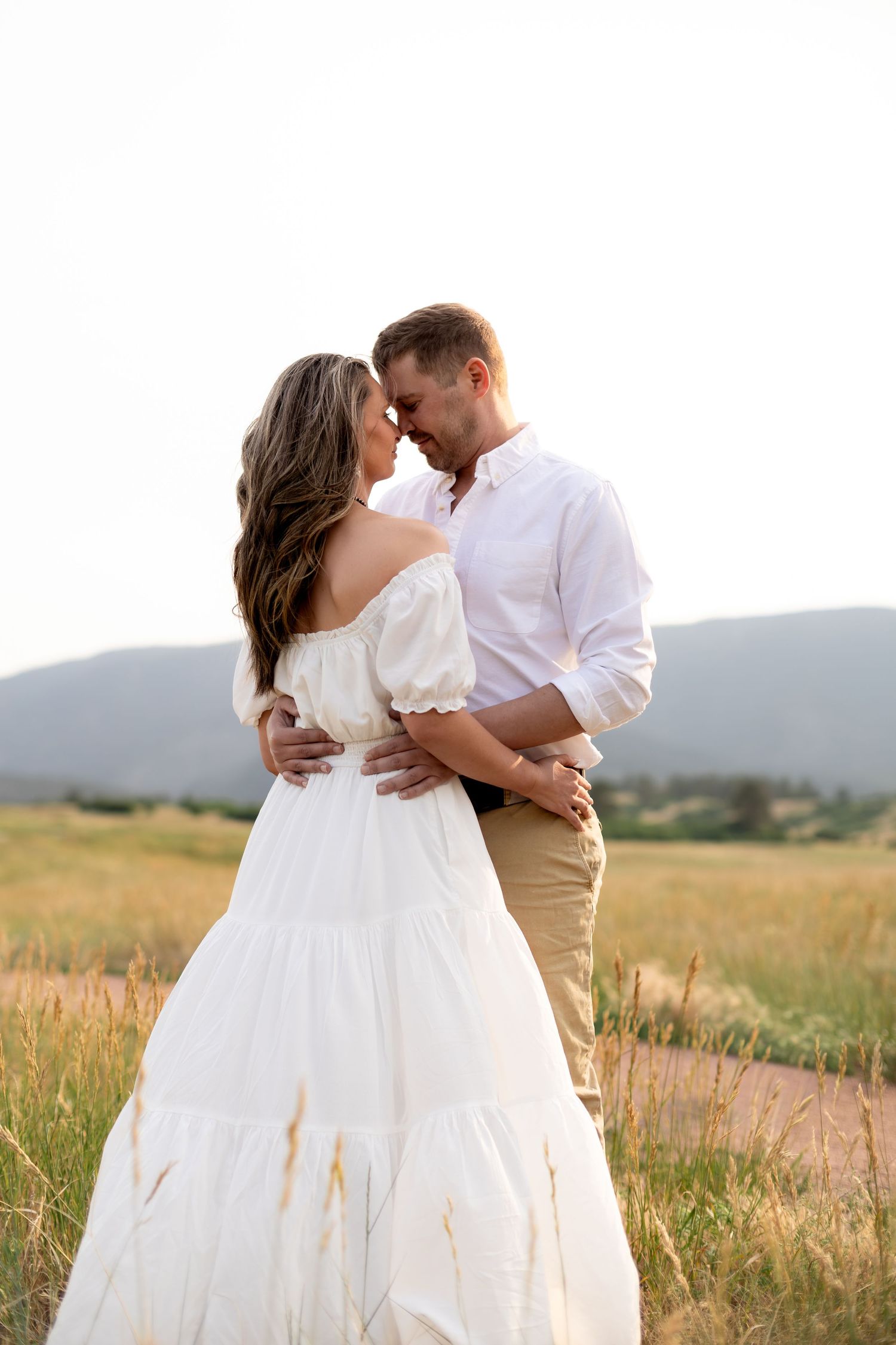 Couple embracing in golden field, woman in white off-shoulder dress, man in white shirt, mountains in background.