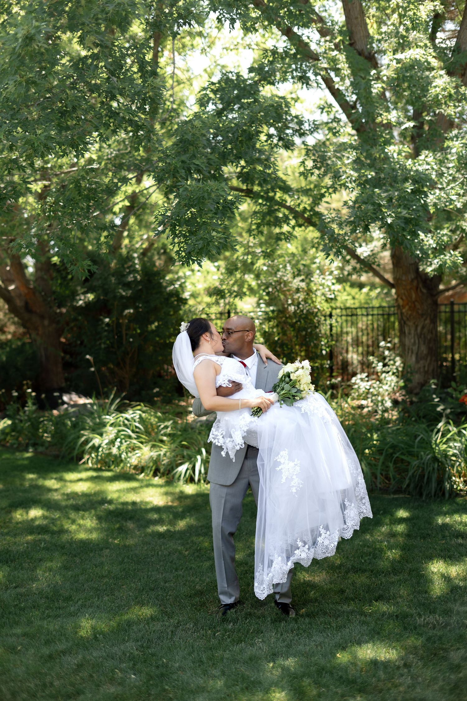 Groom lifts bride in white gown for a kiss in a lush green garden during an outdoor wedding ceremony.