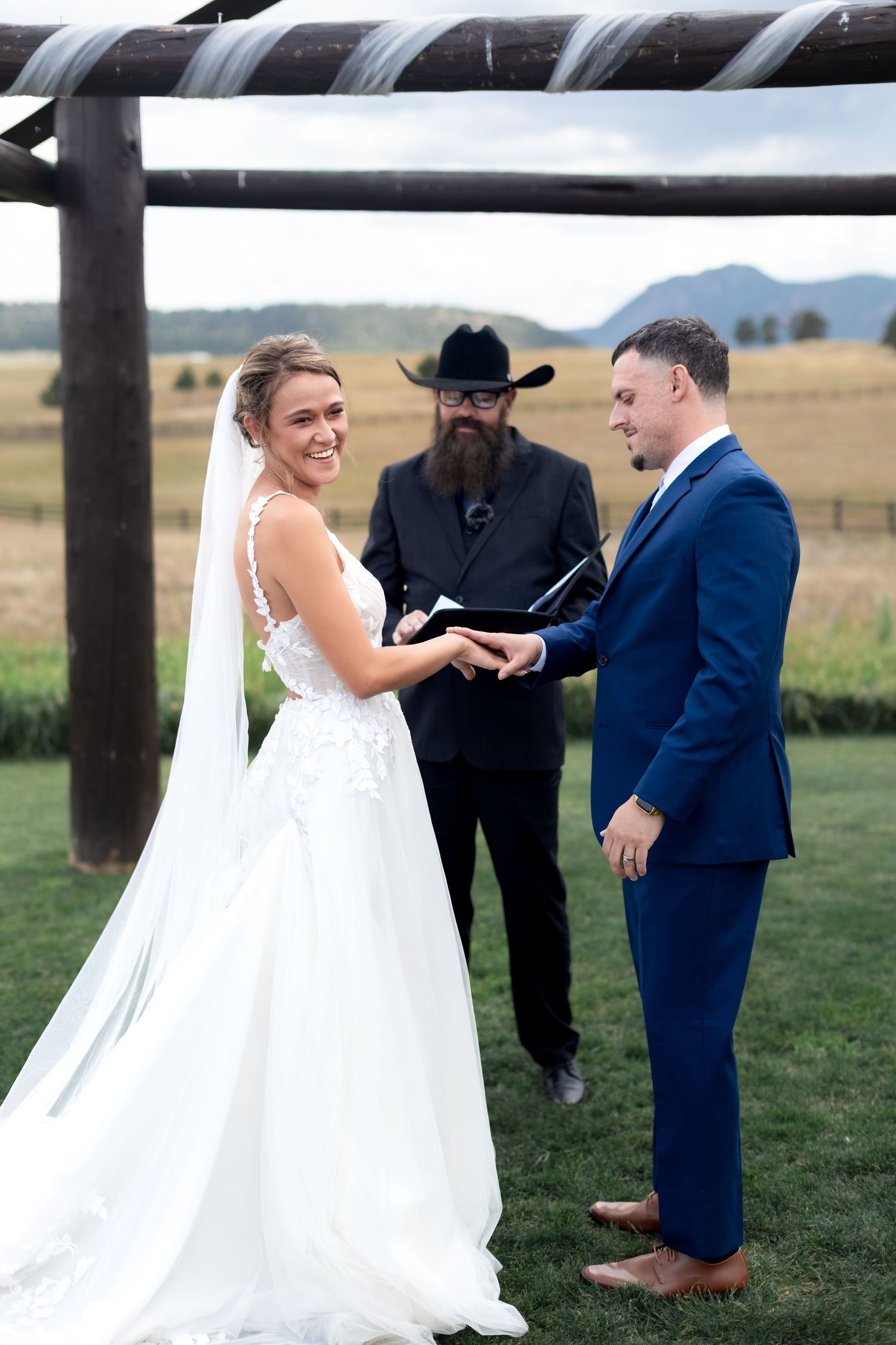 Bride and groom exchange vows outdoors at a rustic ranch wedding with a bearded officiant in a black cowboy hat.
